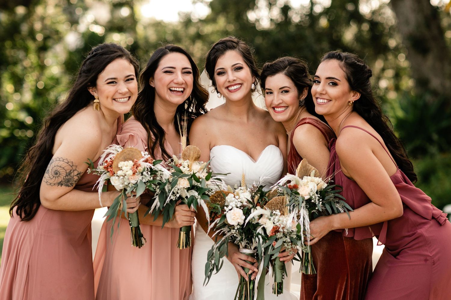 Bride in white strapless gown poses with four bridesmaids in pink and mauve dresses holding floral bouquets outdoors.