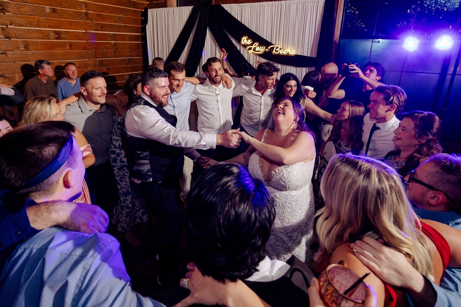 Bride and groom dancing joyfully with wedding guests on a crowded dance floor at a rustic barn reception.