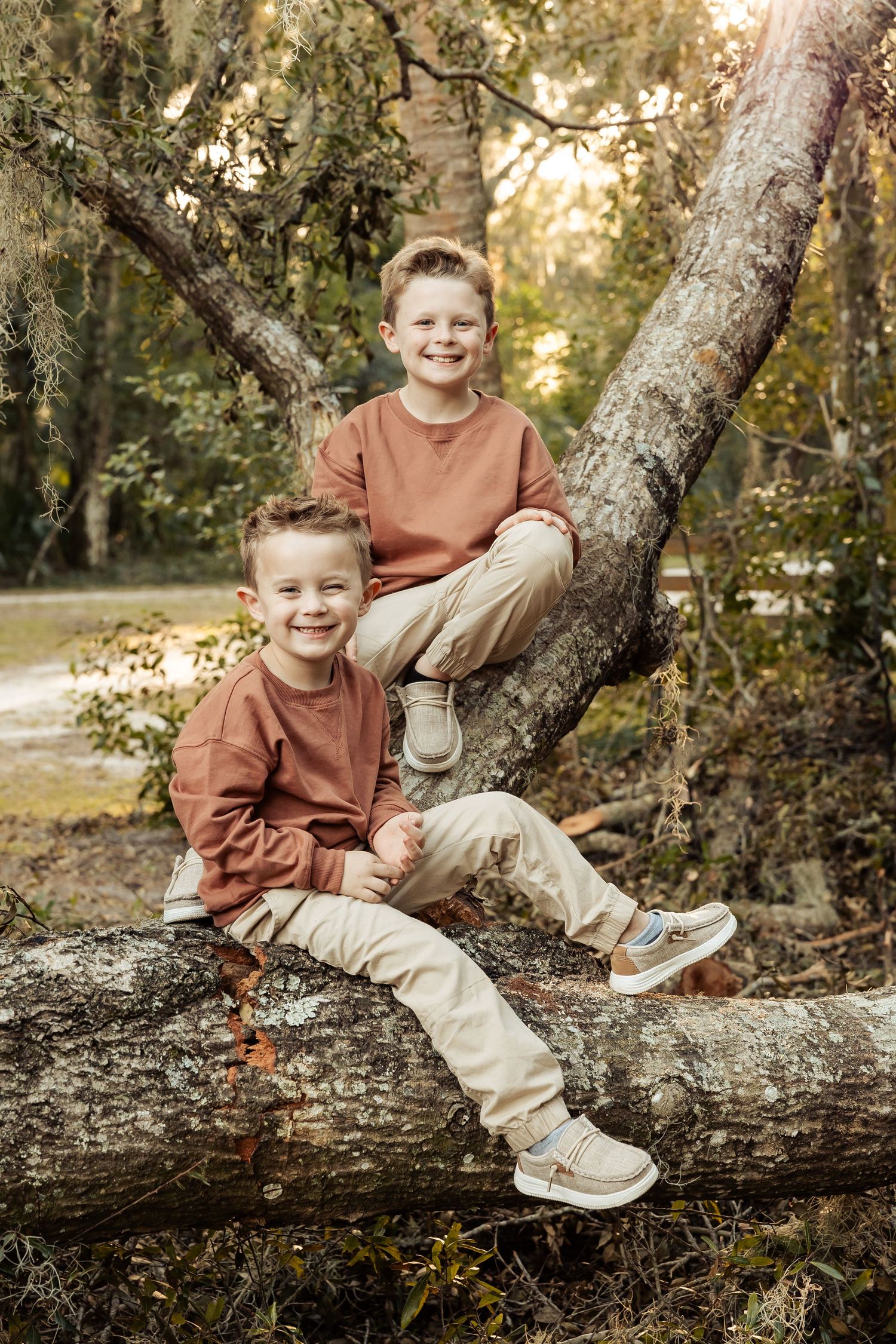 Two smiling boys in matching brown and tan outfits pose on a fallen tree in a wooded park.