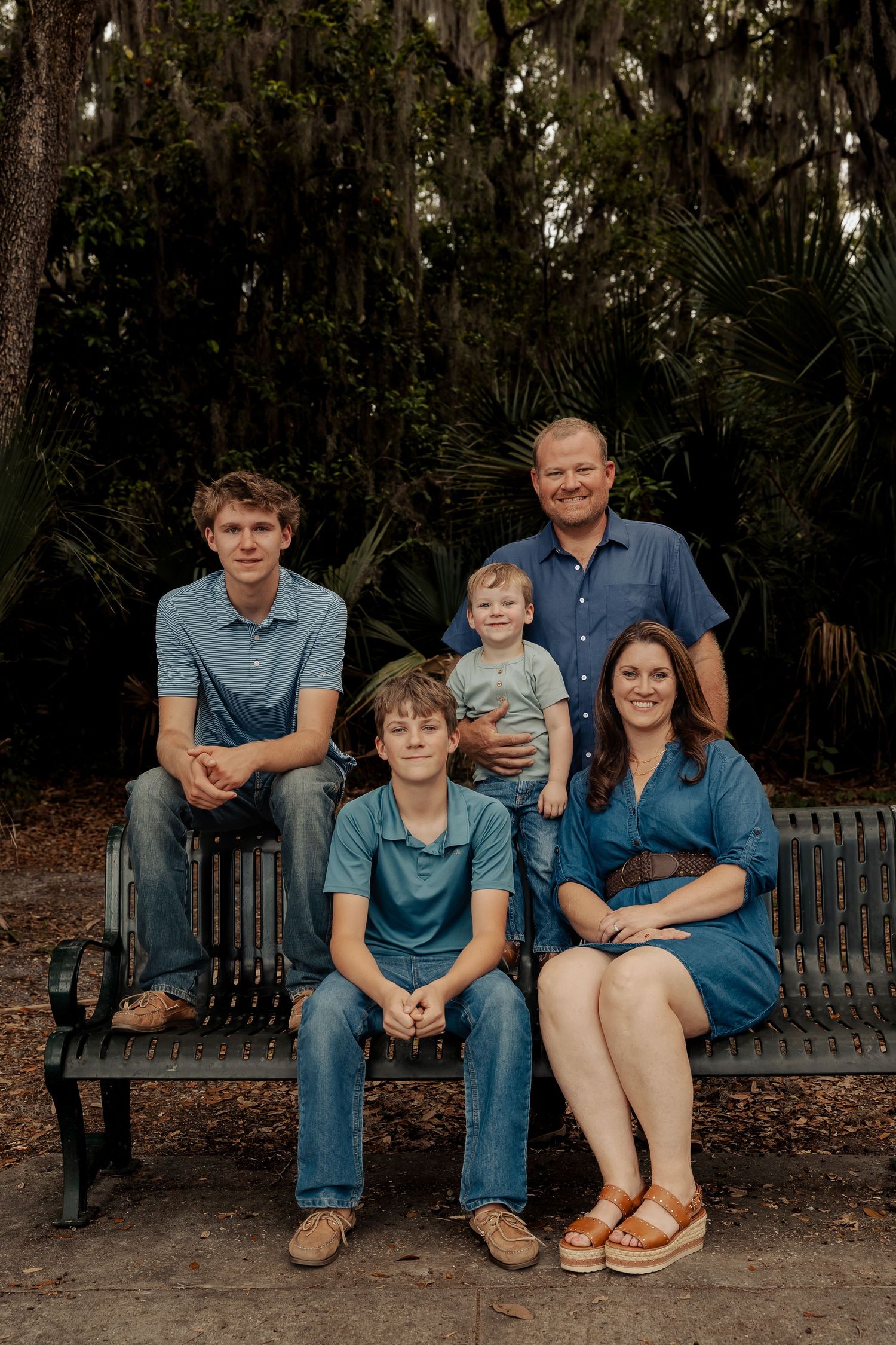 Family of five posing on a park bench surrounded by lush trees, wearing coordinating shades of blue.