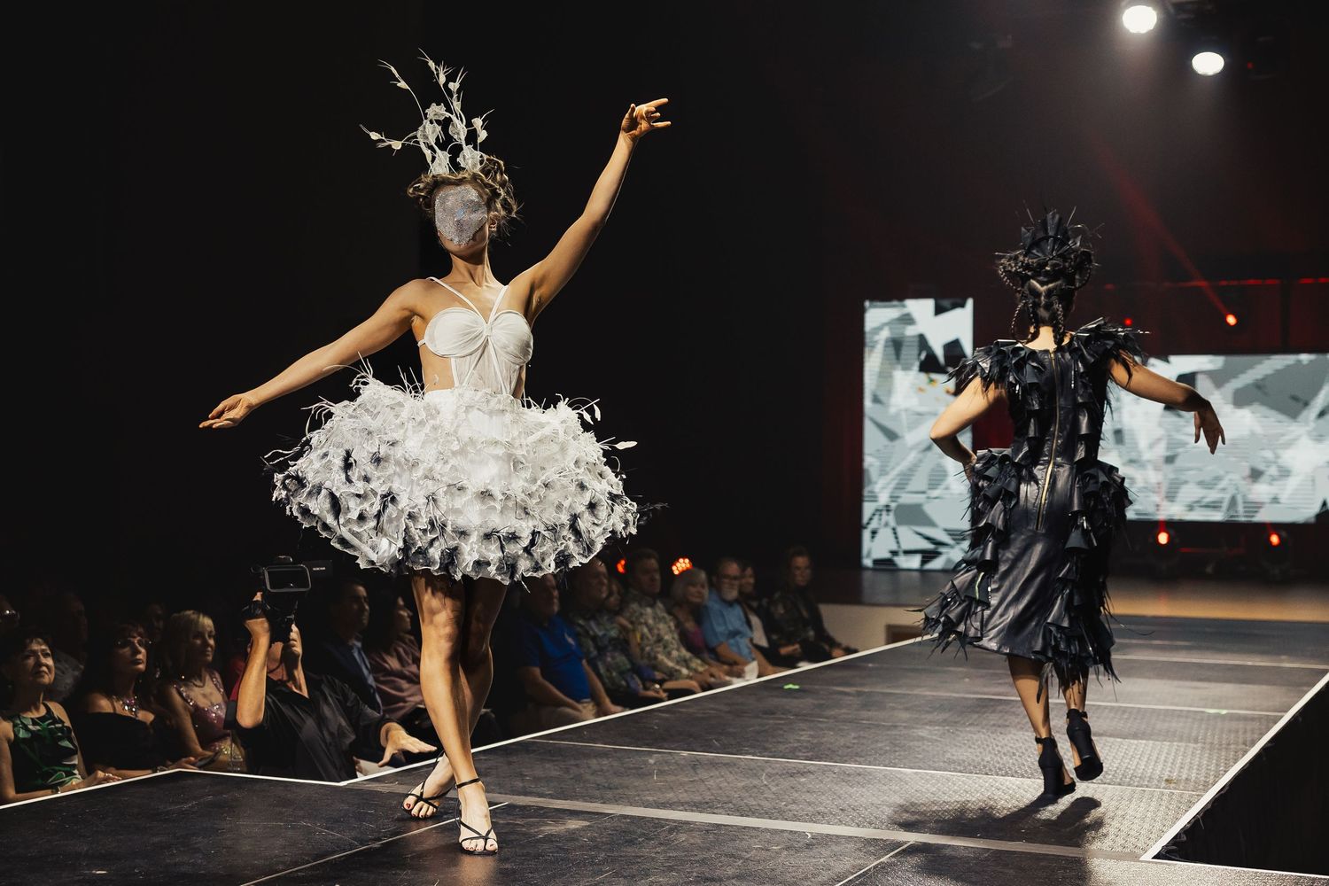Two models in contrasting white feathered and black dramatic costumes walk together on fashion runway.