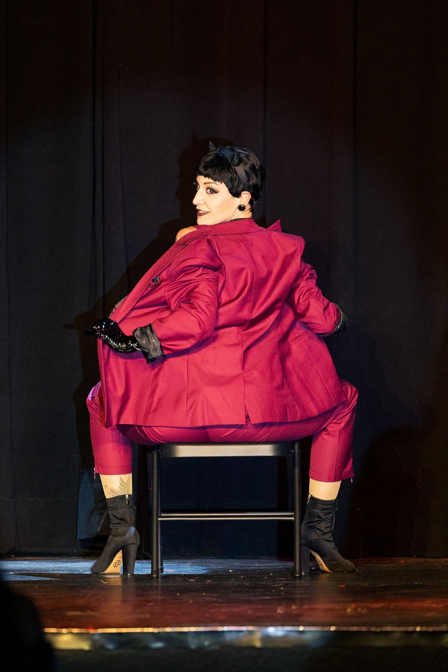 A dark-haired performer in a red suit sits posed on a chair on a darkly lit stage.