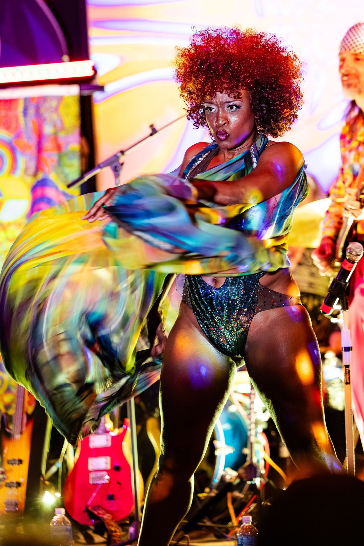 Dynamic dancer with red afro in sparkling costume twirling colorful fabric on a brightly lit stage.