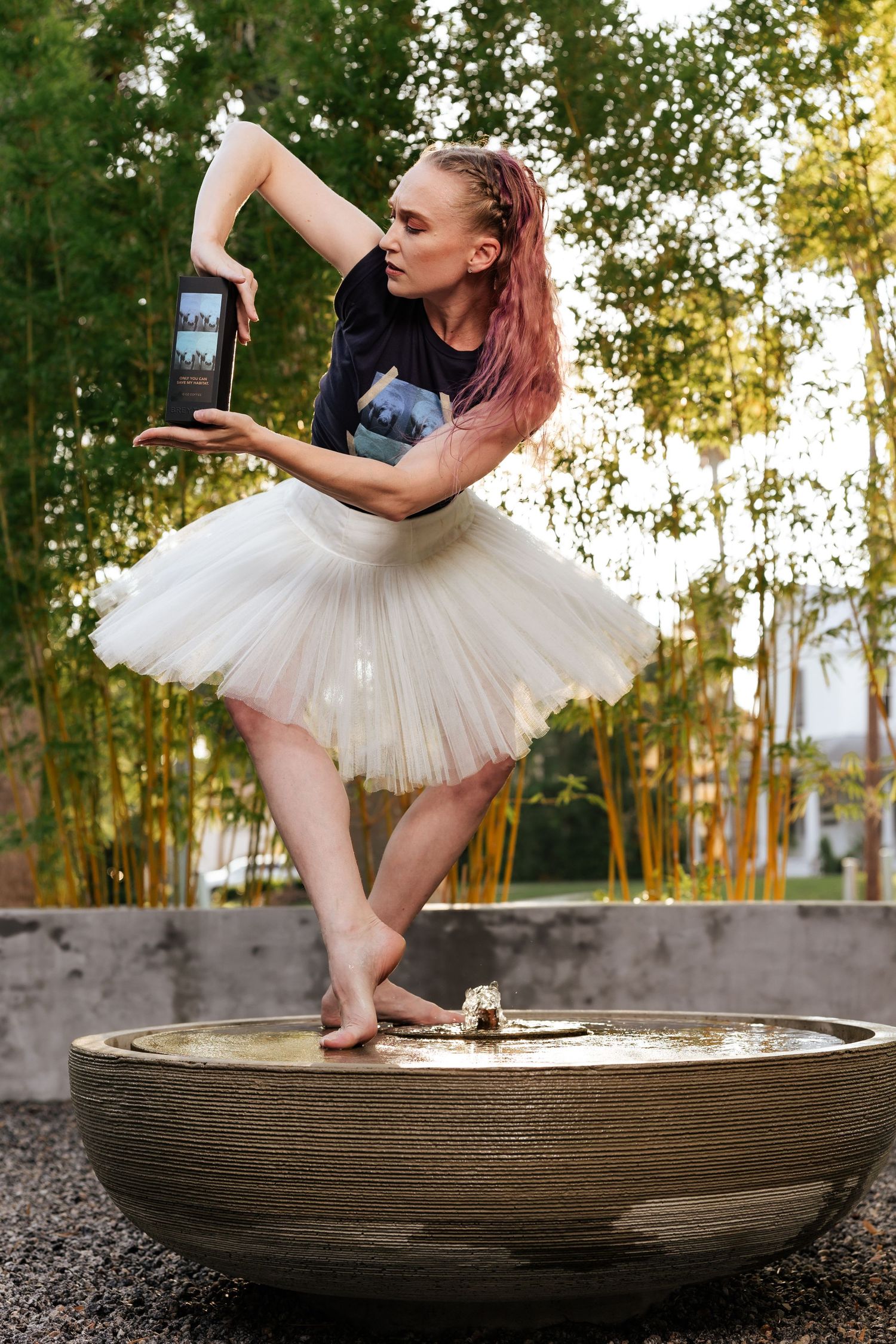 Pink-haired dancer in white tutu performs a ballet pose in an outdoor fountain with bamboo background.