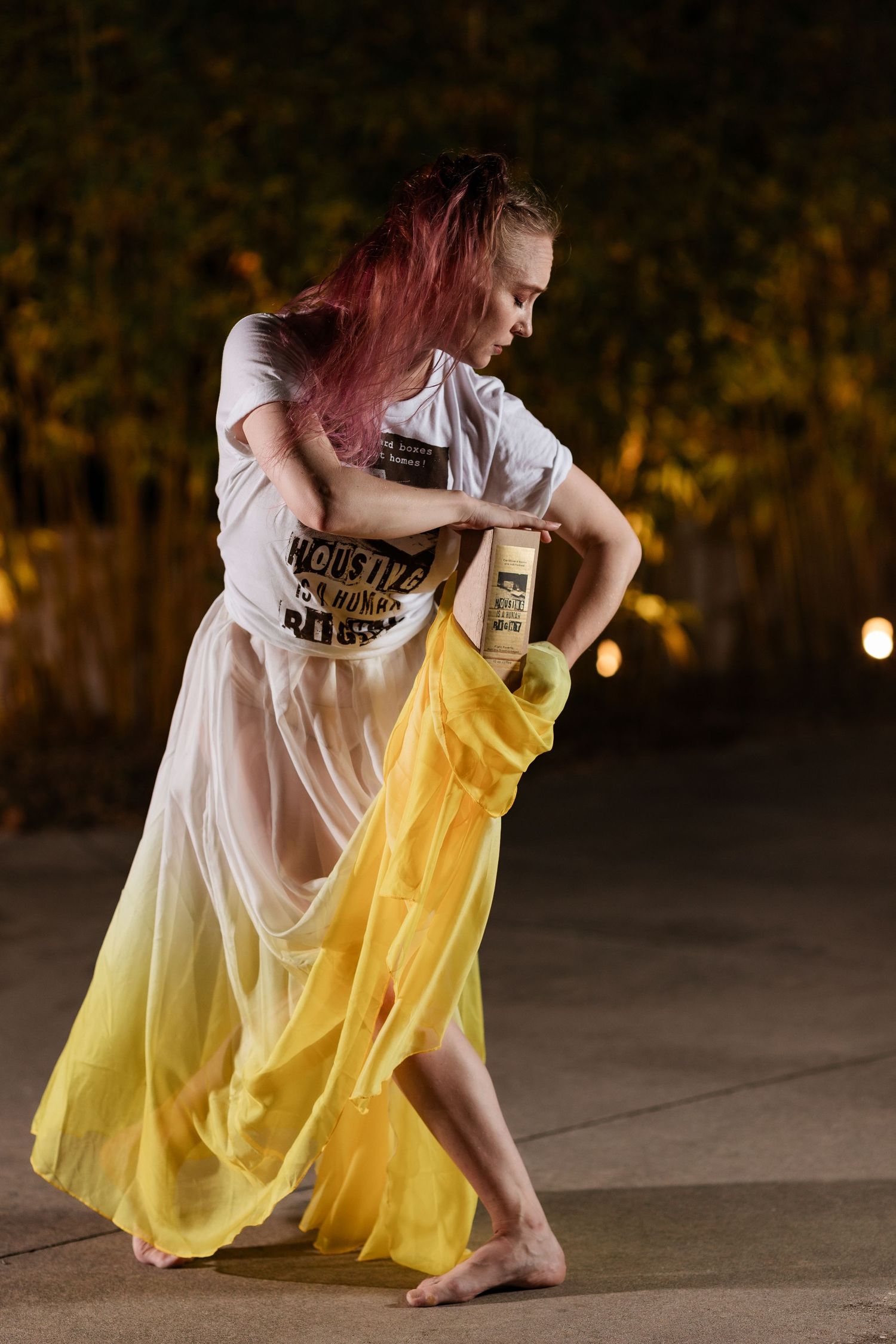 A dancer with pink hair performs barefoot in a sheer yellow skirt holding a bottle outdoors at night.