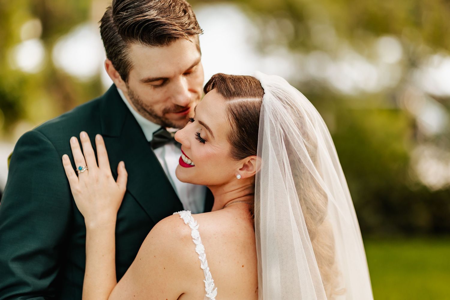 Groom in dark suit tenderly embraces bride in lace gown with veil in soft outdoor light.