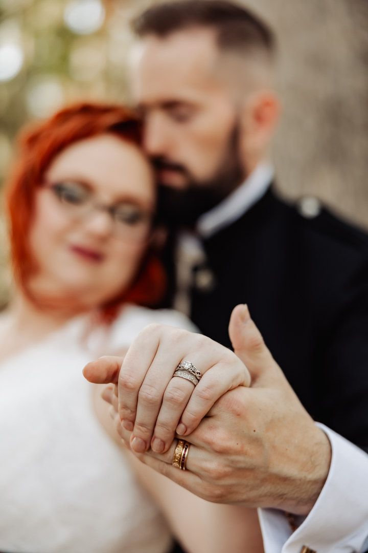 Close-up of bride and groom holding hands displaying gold wedding rings with blurred couple in background.