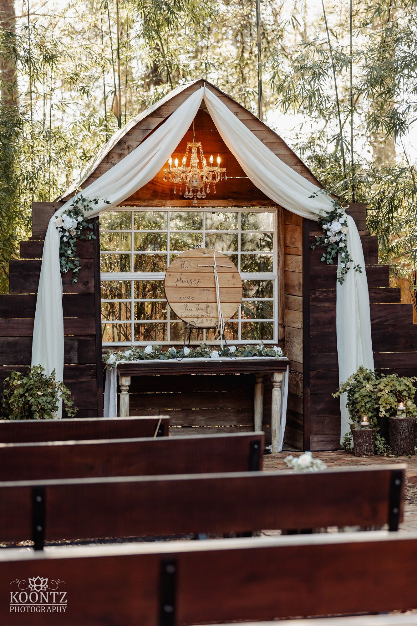 Rustic outdoor wedding ceremony altar with white draping, chandelier, and wooden chapel structure surrounded by trees.