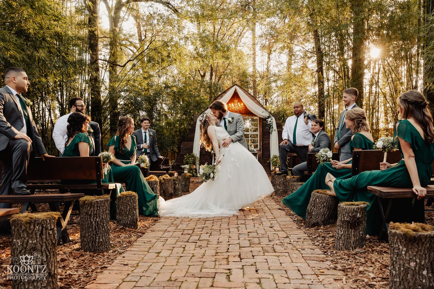 Bride walks down brick aisle at outdoor woodland wedding ceremony with green bridesmaid dresses and log seating.