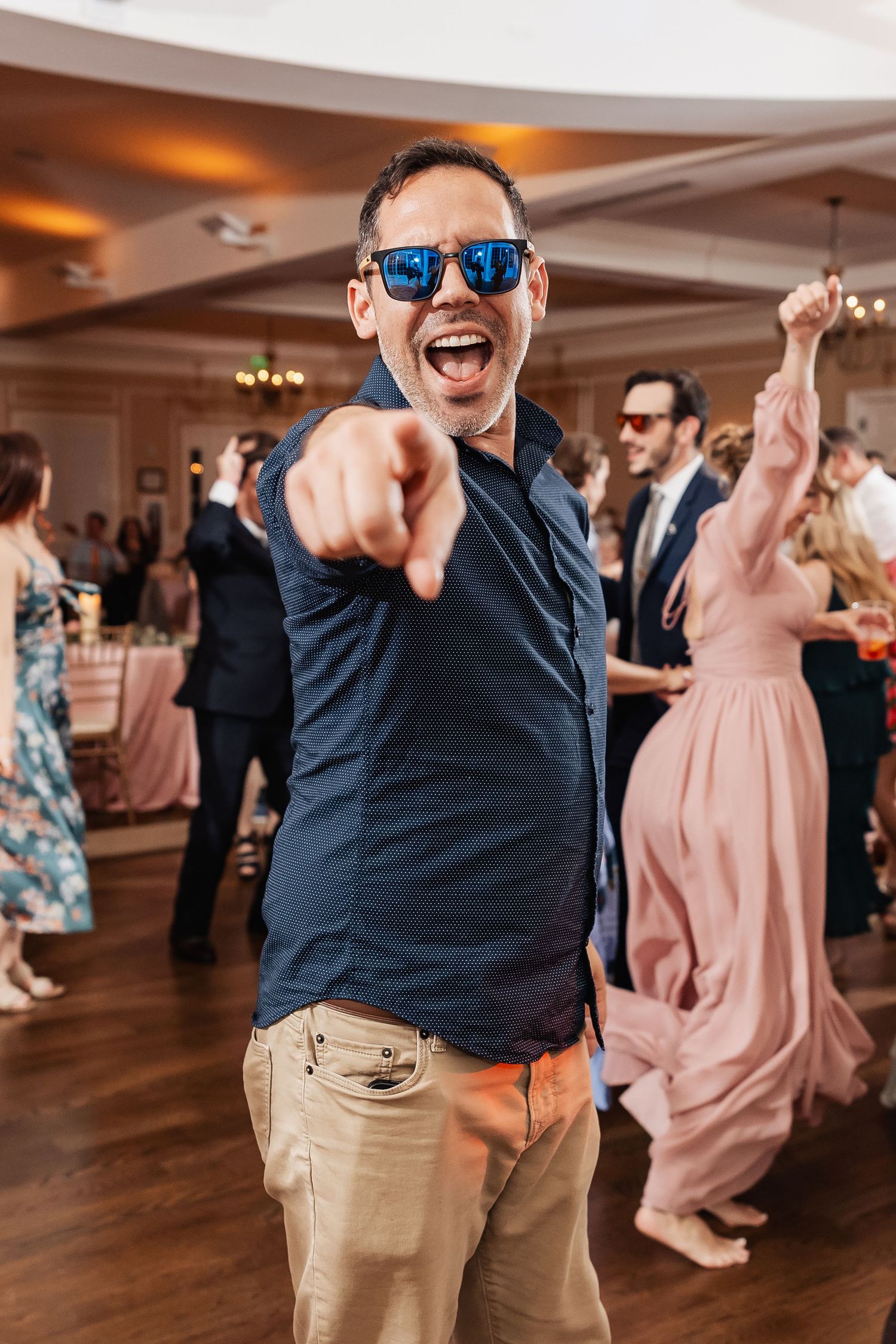 Excited man in sunglasses pointing at camera while dancing at a wedding reception.
