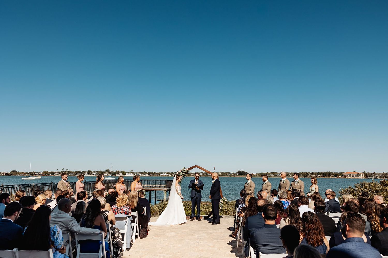 Outdoor waterfront wedding ceremony with guests seated on both sides as couple stands at wooden arch.
