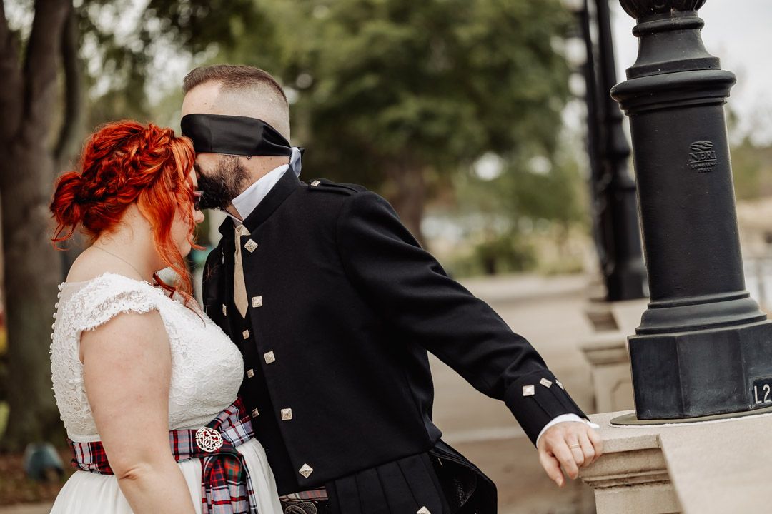 Wedding couple in Scottish attire walking together on a path with lamp posts.