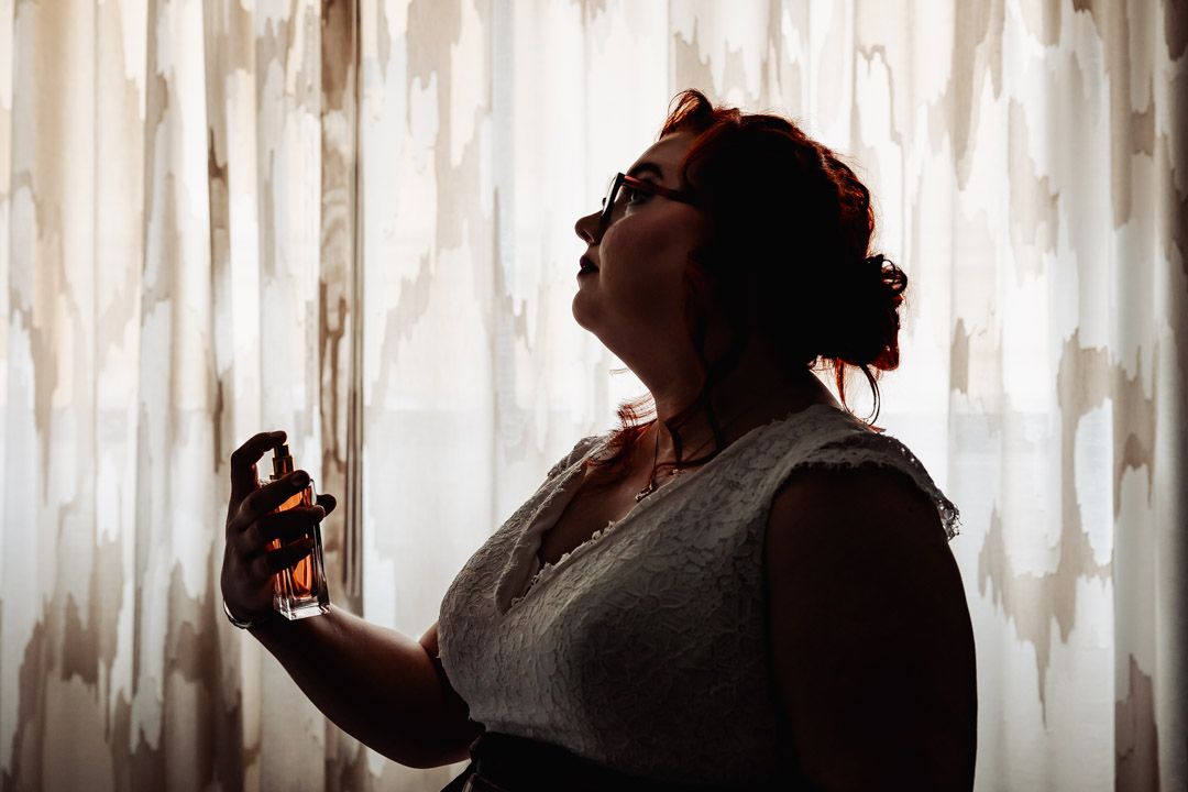 Silhouette of someone holding a perfume bottle against a backlit white curtained window.