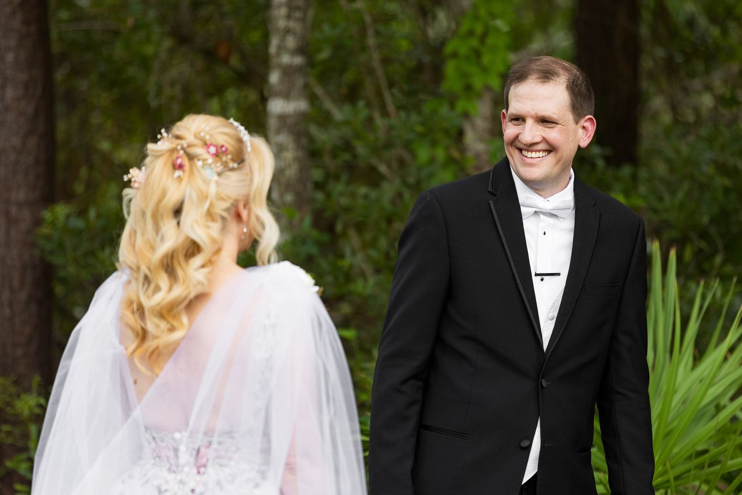 Groom smiling warmly at blonde bride during romantic outdoor first look in lush green garden.