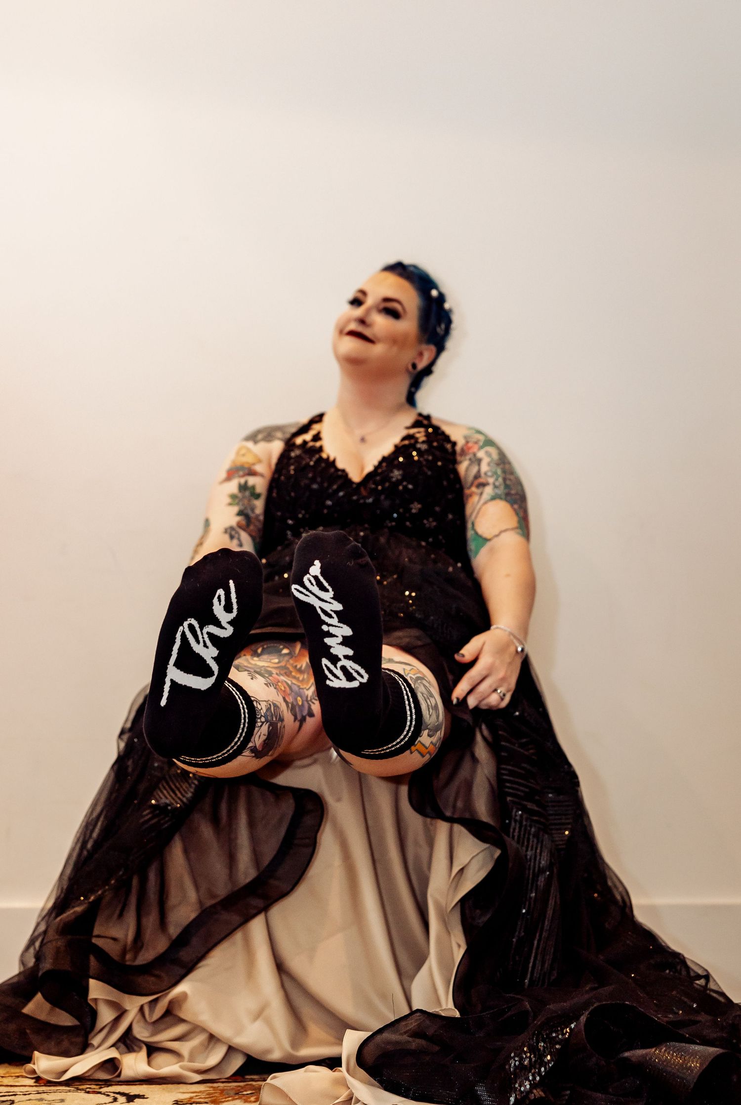 Tattooed bride in black sequin gown showing off The Bride sneakers while seated against white wall.
