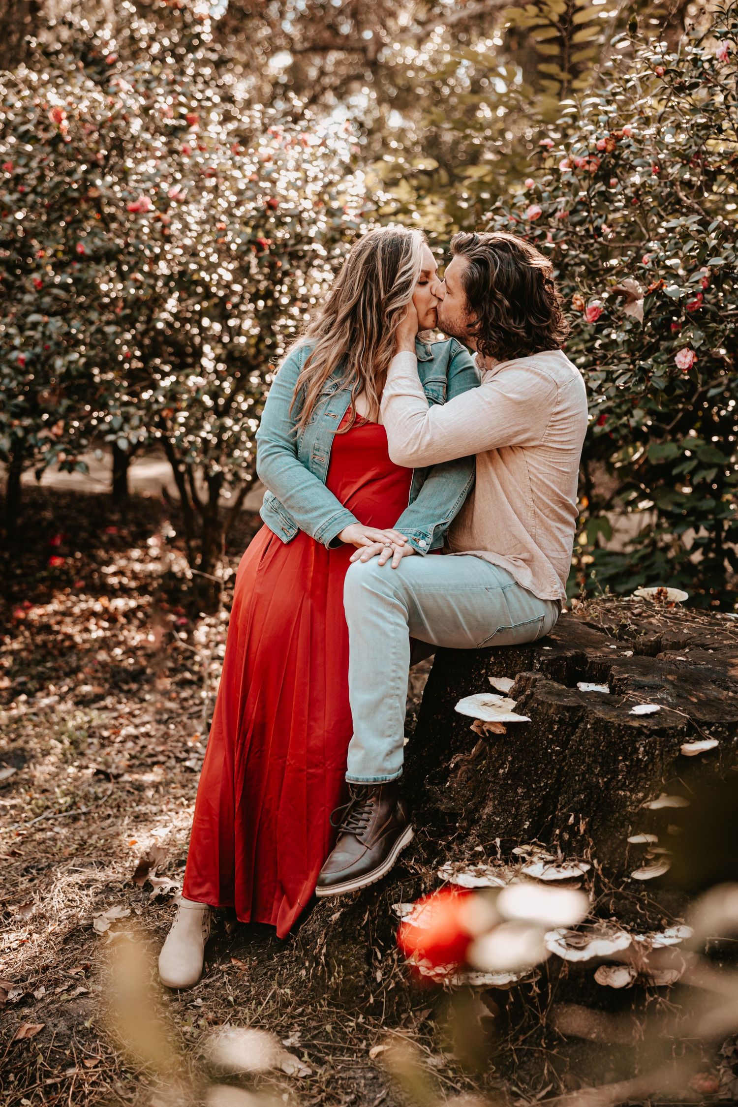 Couple sharing a kiss while sitting on a stone wall surrounded by blooming red camellia trees.
