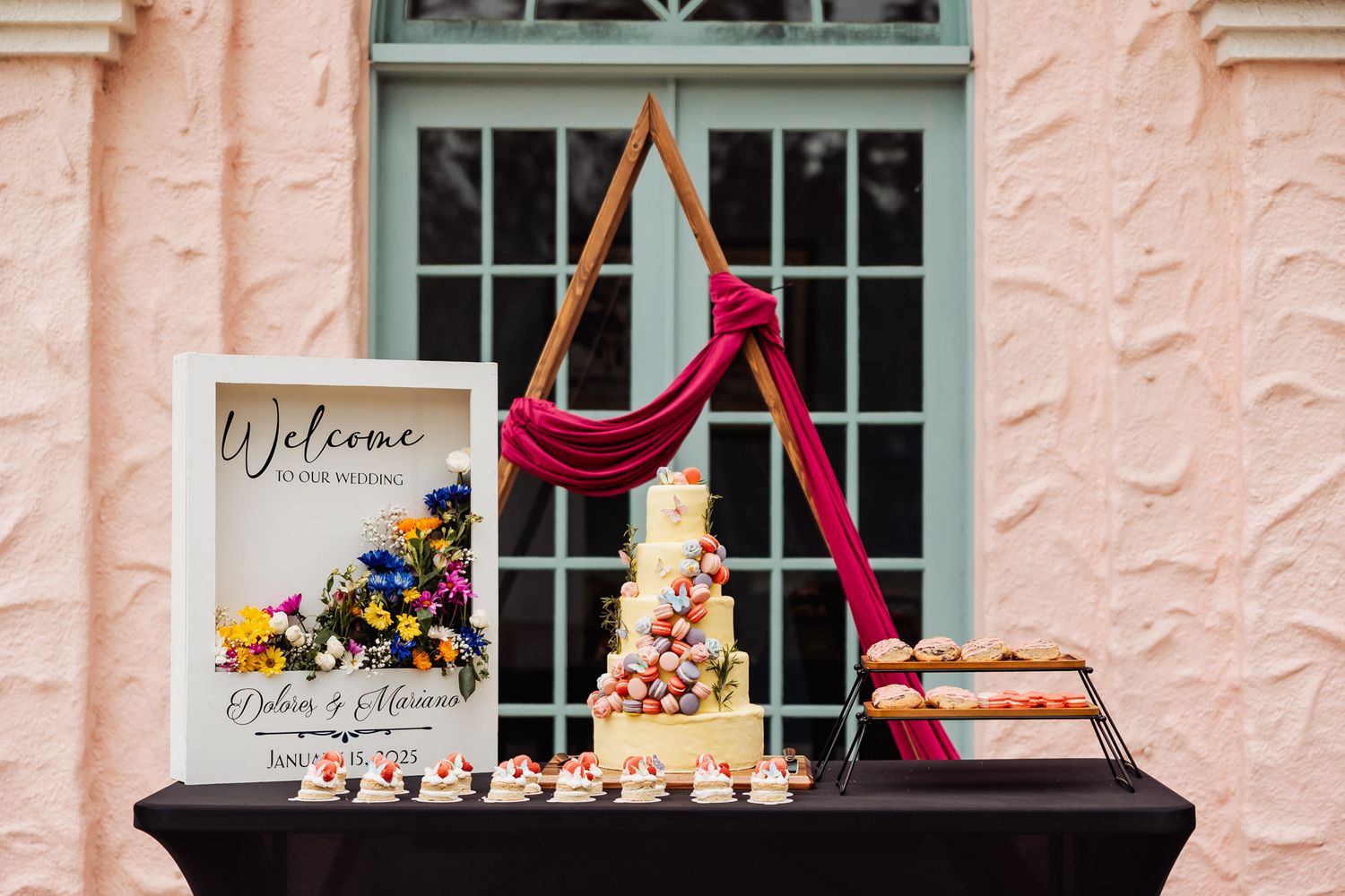 Elegant wedding display featuring a tall tiered cake with burgundy draping and welcome sign against a pink stucco wall.