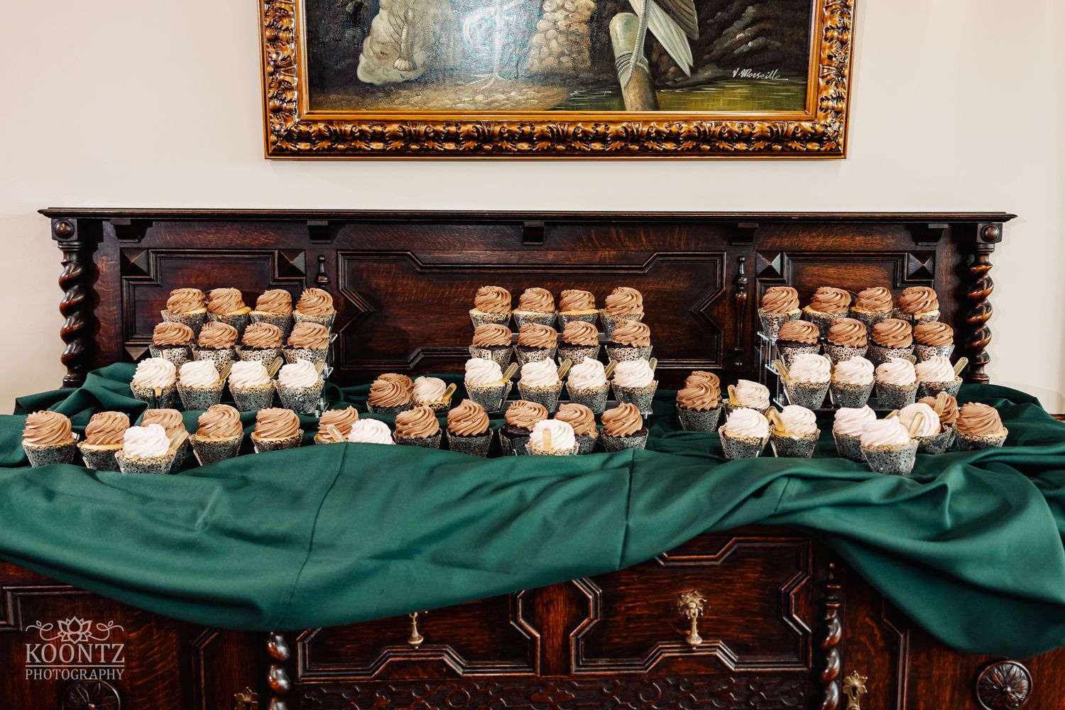 Display of elegant cupcakes arranged on dark wooden buffet with green tablecloth.