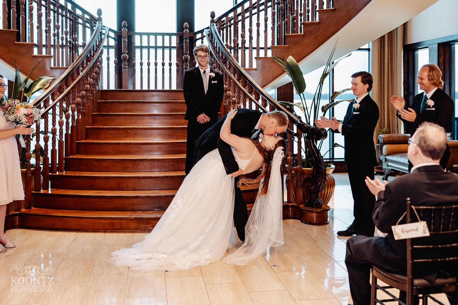 Wedding party and guests gathered around elegant wooden staircase during celebration.