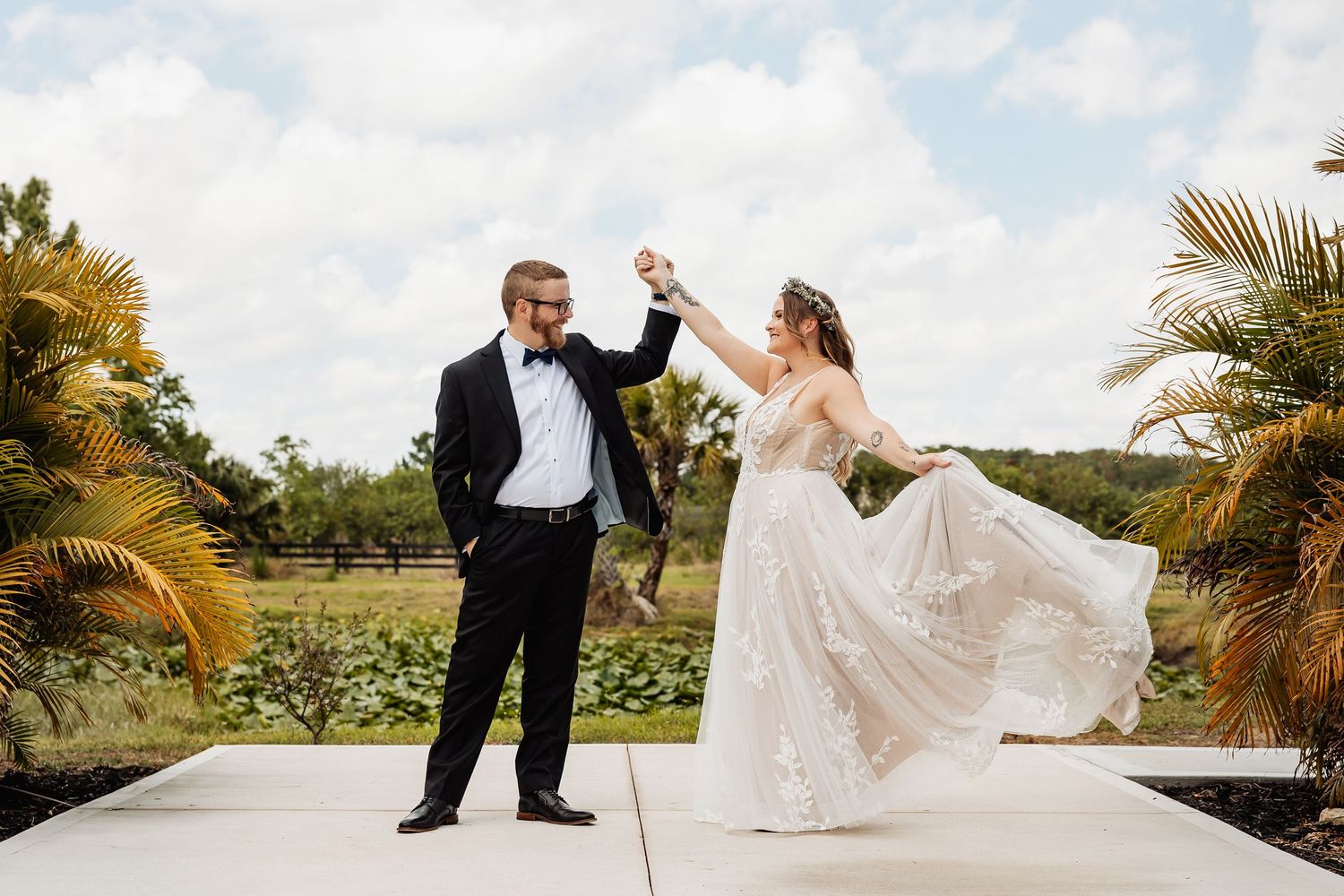 A joyful couple in formal wedding attire dances outdoors on a white stone patio surrounded by tropical palm trees.