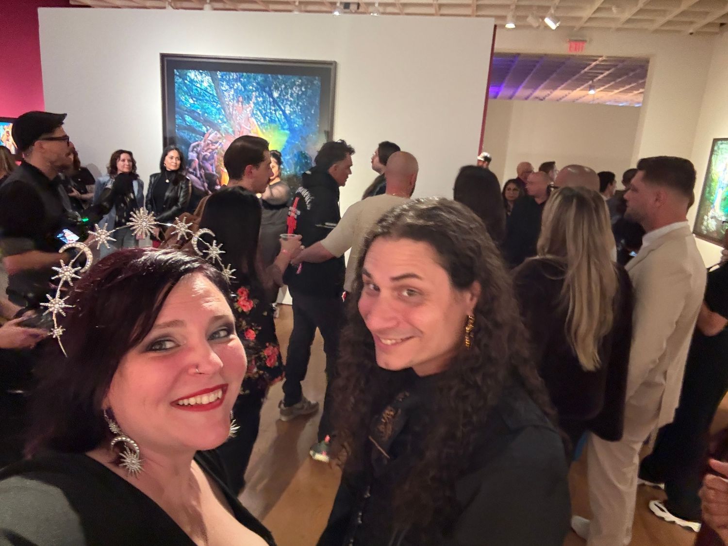 People gathered at an art gallery opening event viewing artwork displayed on white walls.