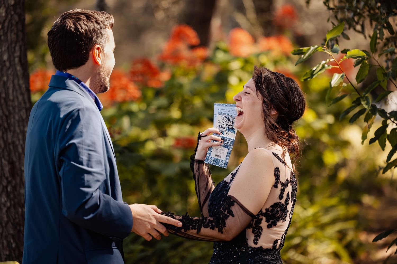 Bride laughing joyfully while reading wedding vows to groom surrounded by vibrant orange flowers.