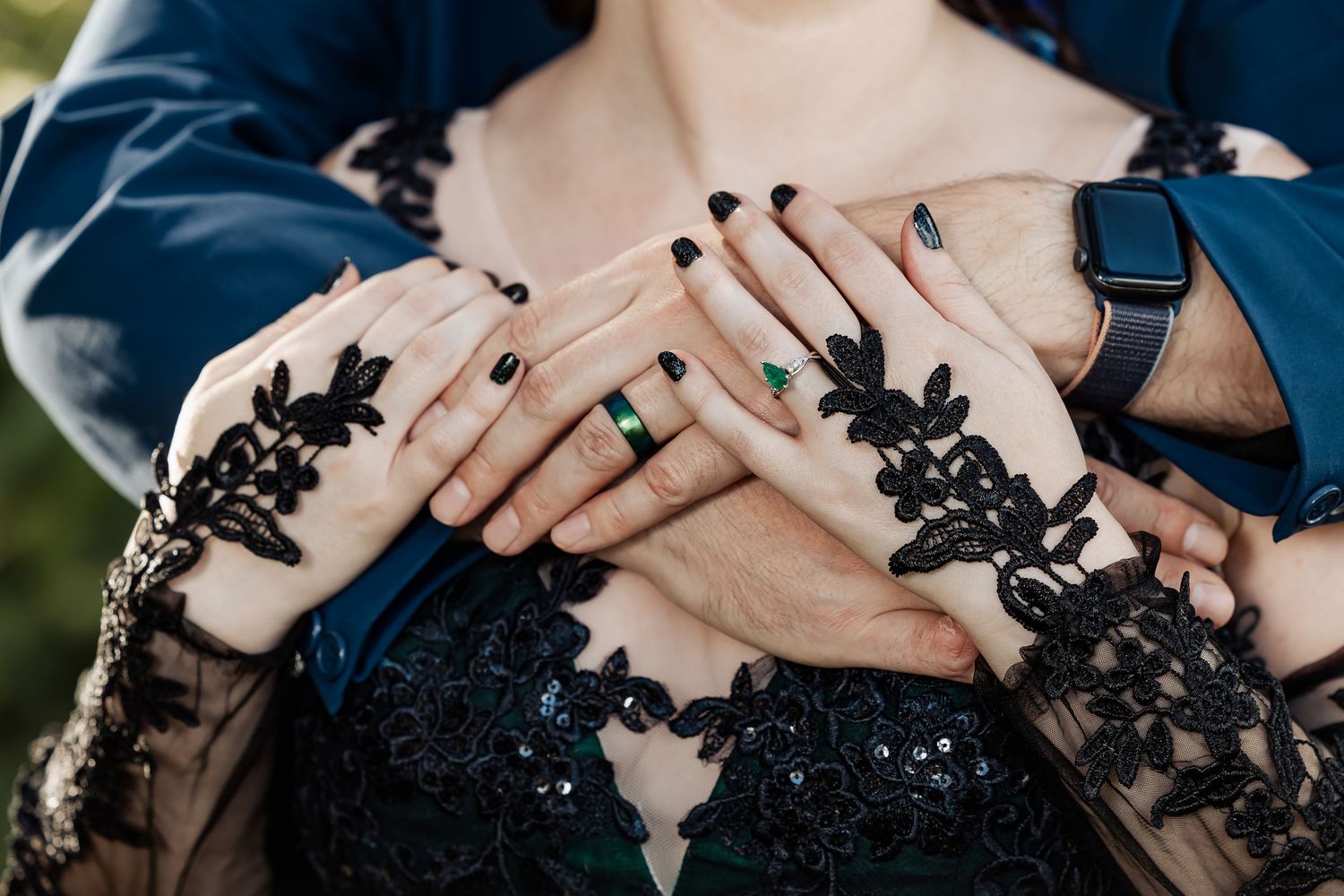 Close-up of bride and groom hands showing black nails, emerald ring, and smartwatch on black lace wedding dress.
