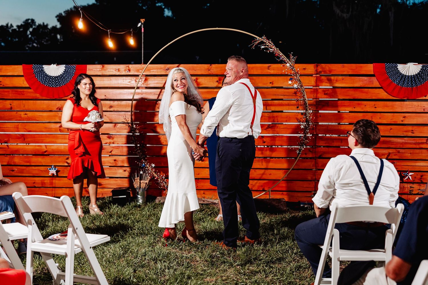 Bride and groom holding hands at outdoor wedding ceremony with circular arch and patriotic decorations on wood wall.