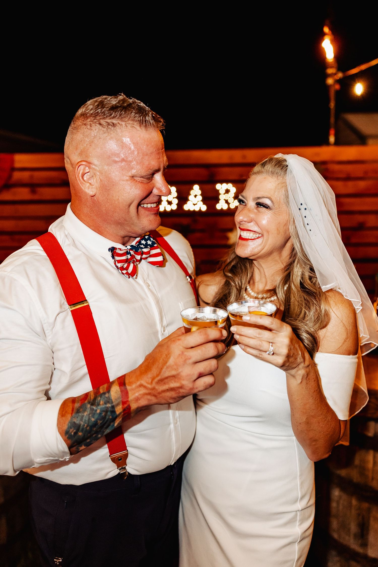 Bride and groom toasting with whiskey glasses at outdoor wedding reception near illuminated bar sign.