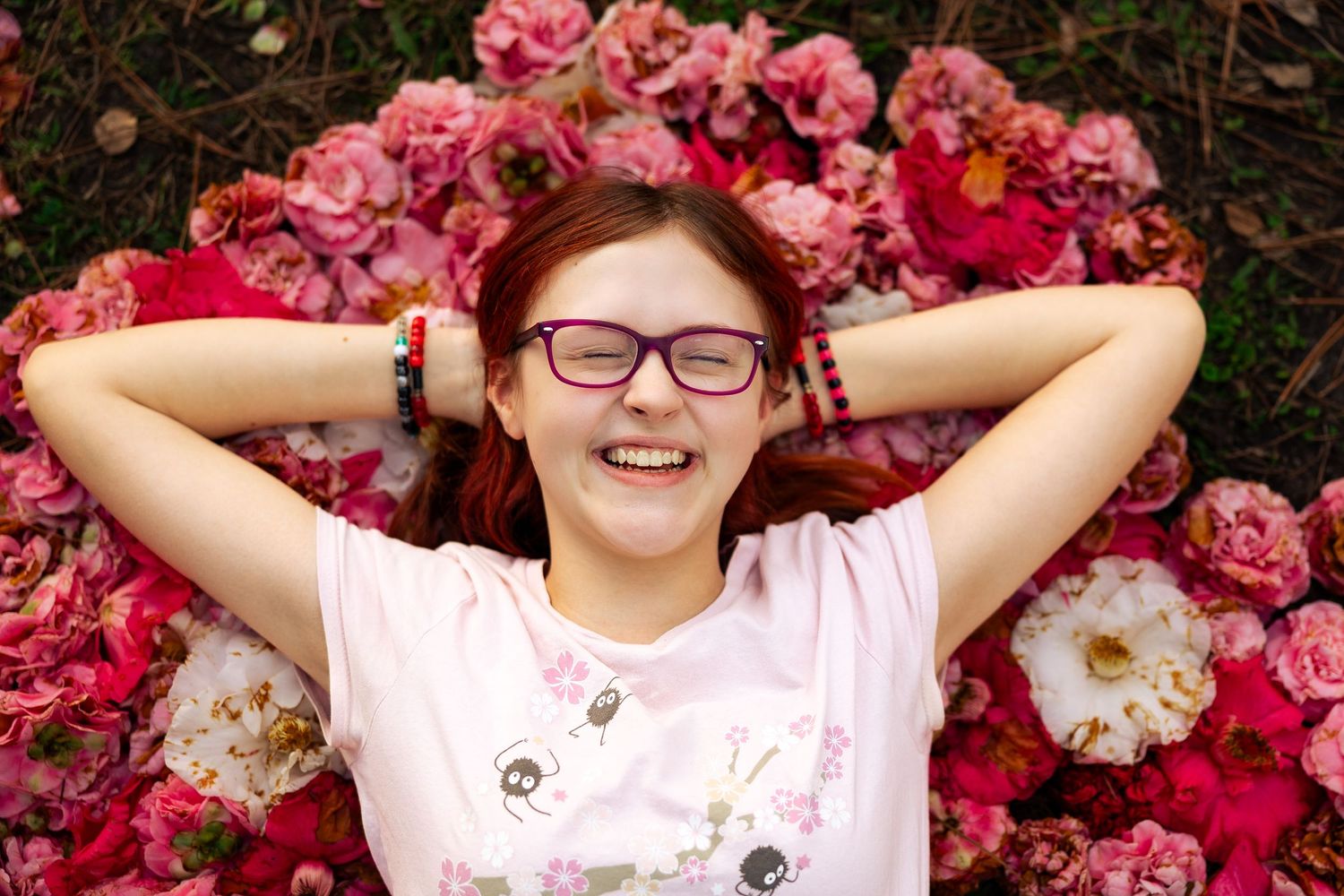 Smiling teenage girl with red hair and glasses lying among pink and white flowers outdoors.