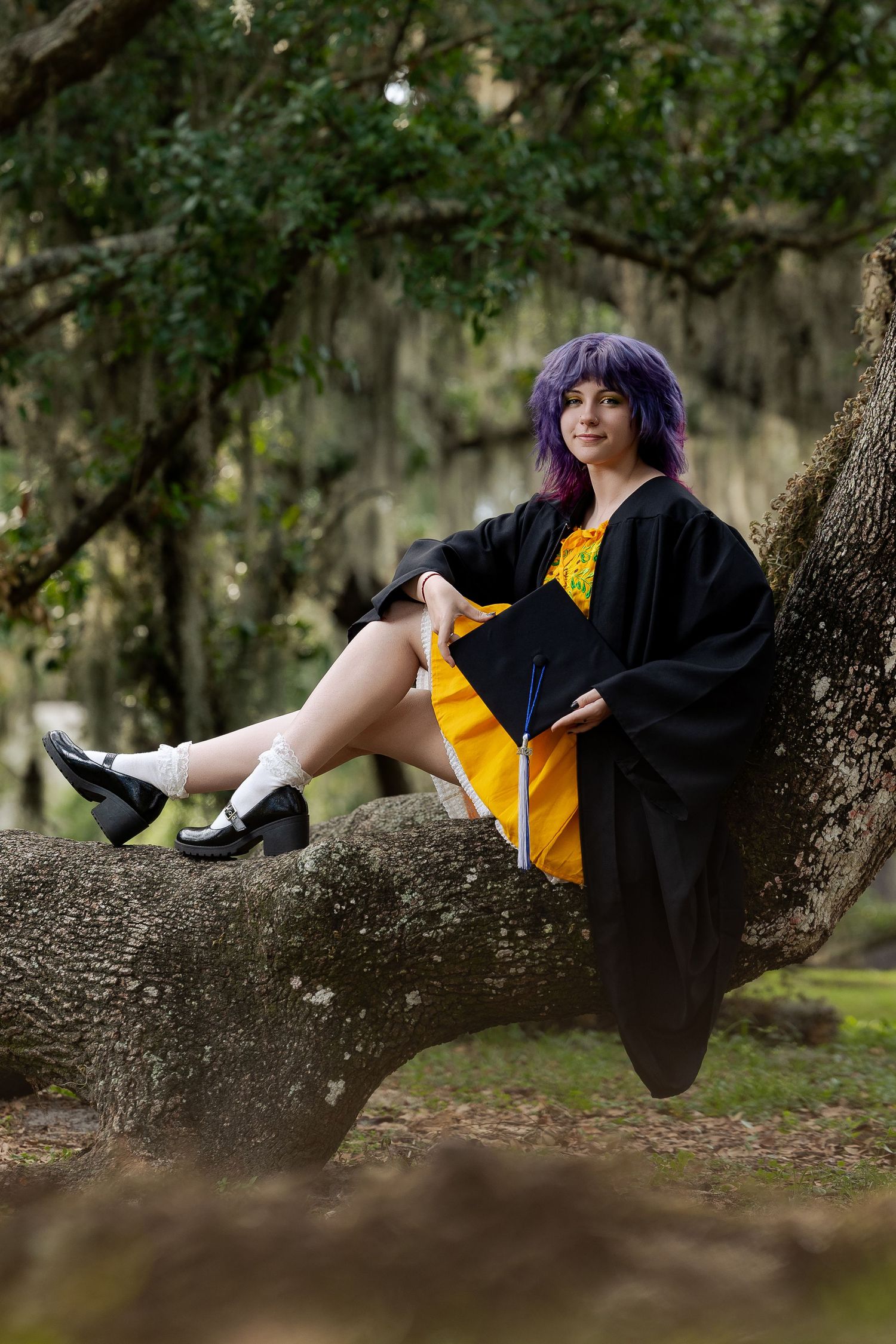 A young woman in graduation gown and cap sits on a tree branch, wearing a yellow dress and purple hair outdoors.