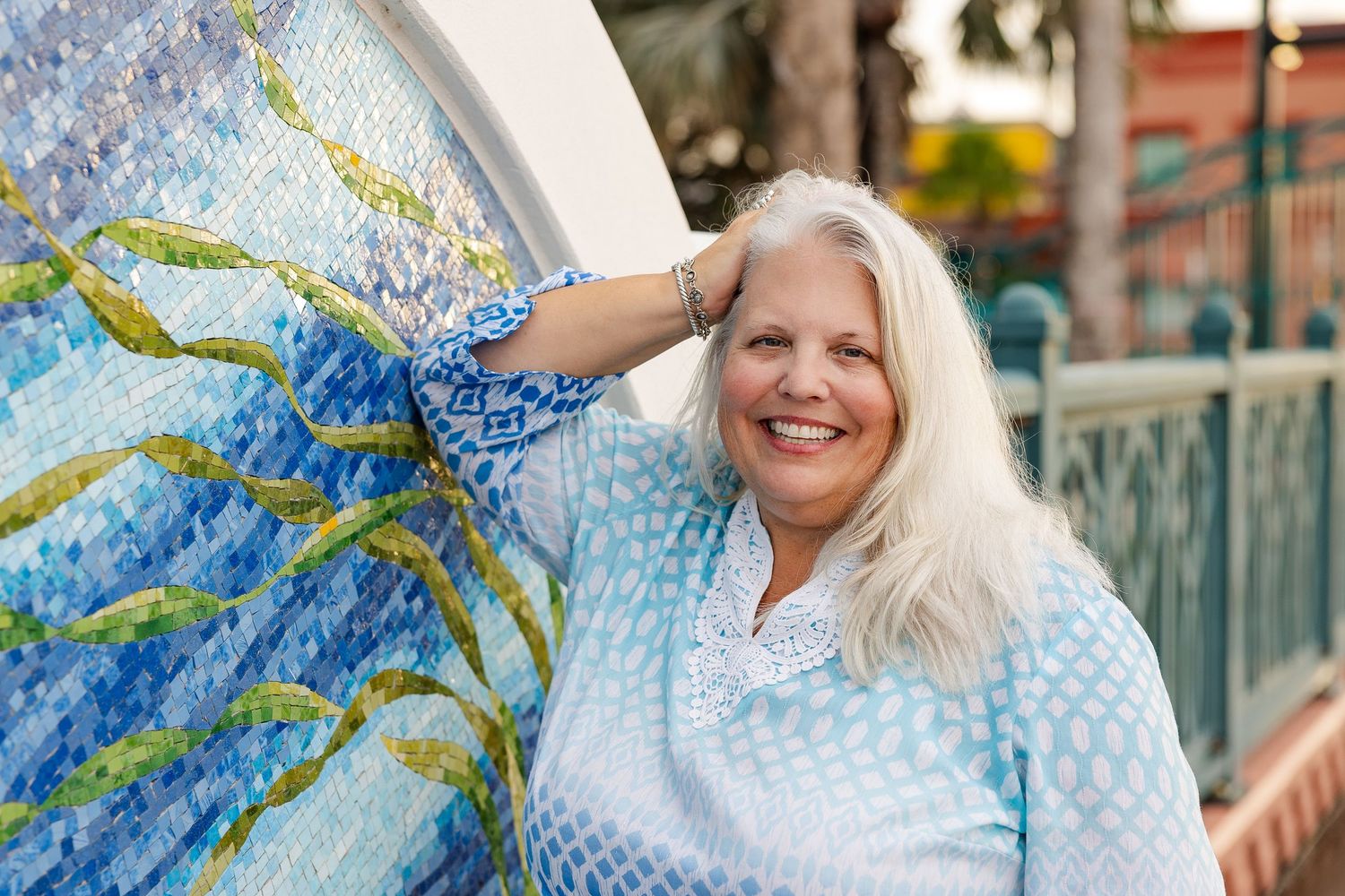 Smiling woman with white hair in blue top leaning against colorful mosaic mural outdoors.