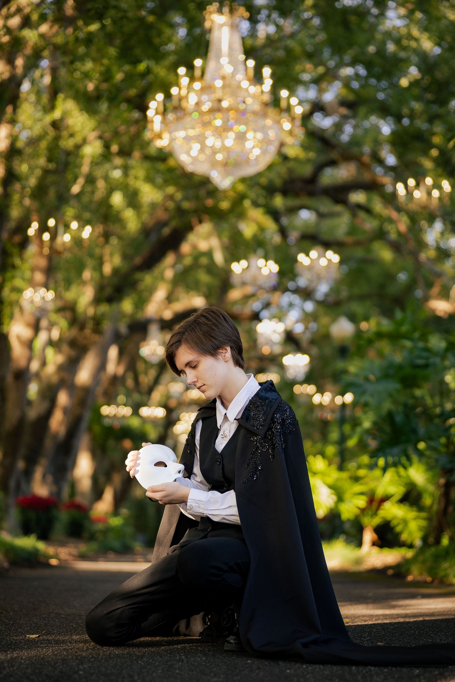 Person in black cape kneeling outdoors holding white masquerade mask with chandelier backdrop.