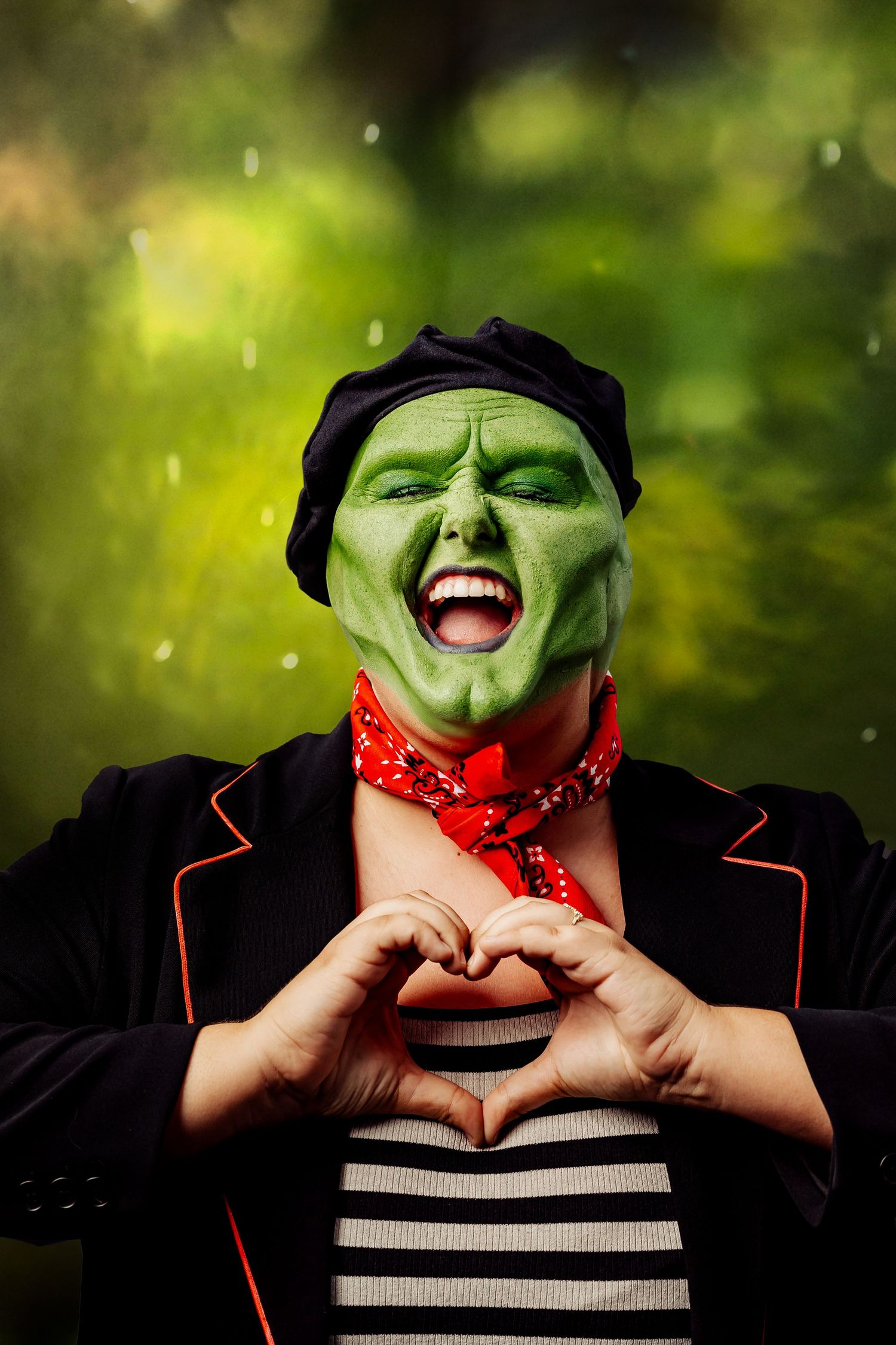Person in green face paint and black beret costume inspired by The Mask, making a heart shape with hands outdoors.