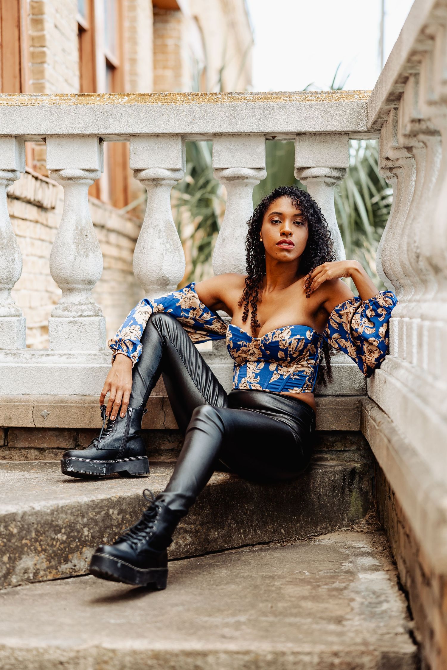 Young woman in blue floral crop top and black leather pants poses on stone steps outdoors.
