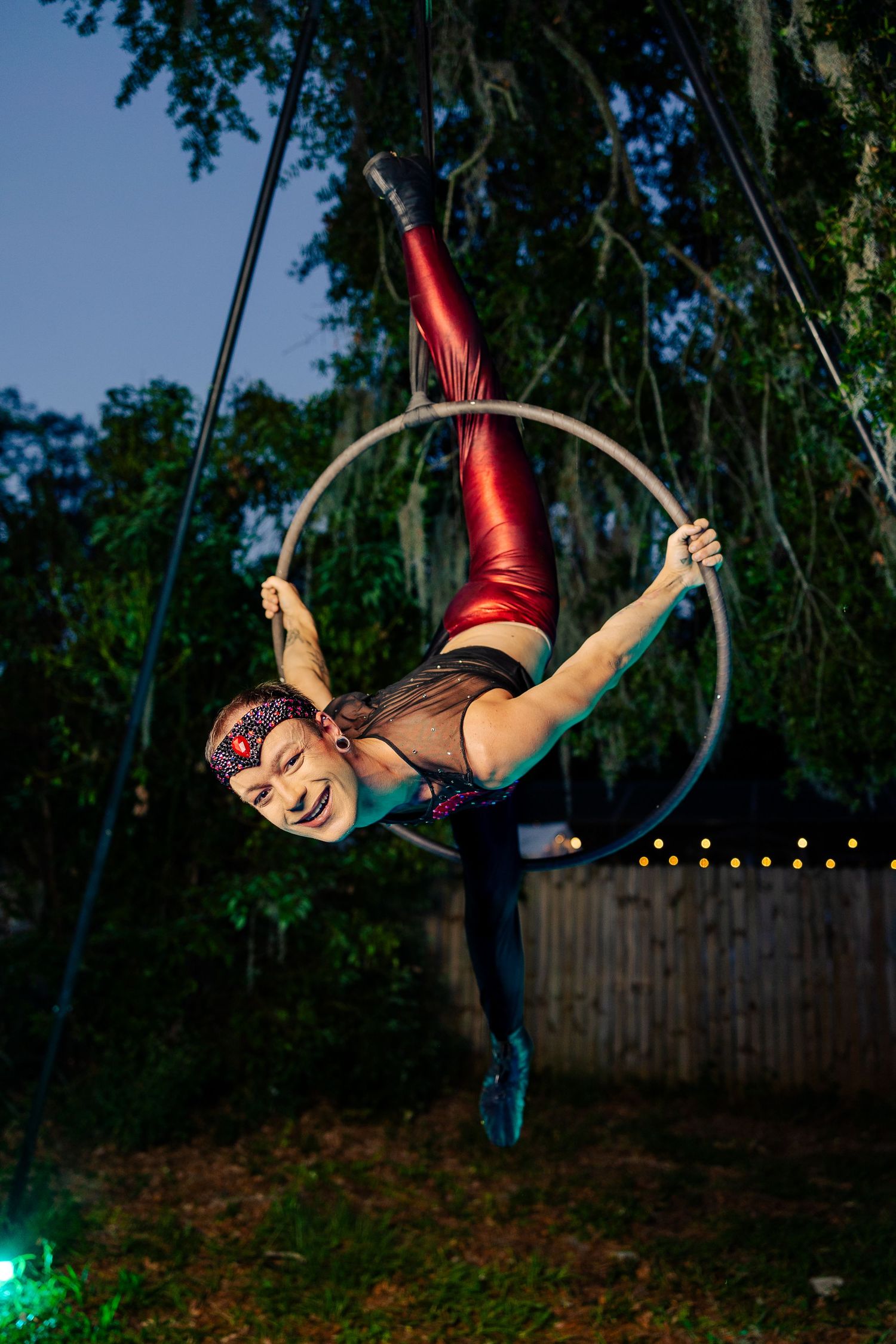 Female aerial hoop artist in red metallic leggings performing dynamic pose on lyra ring outdoors at night.