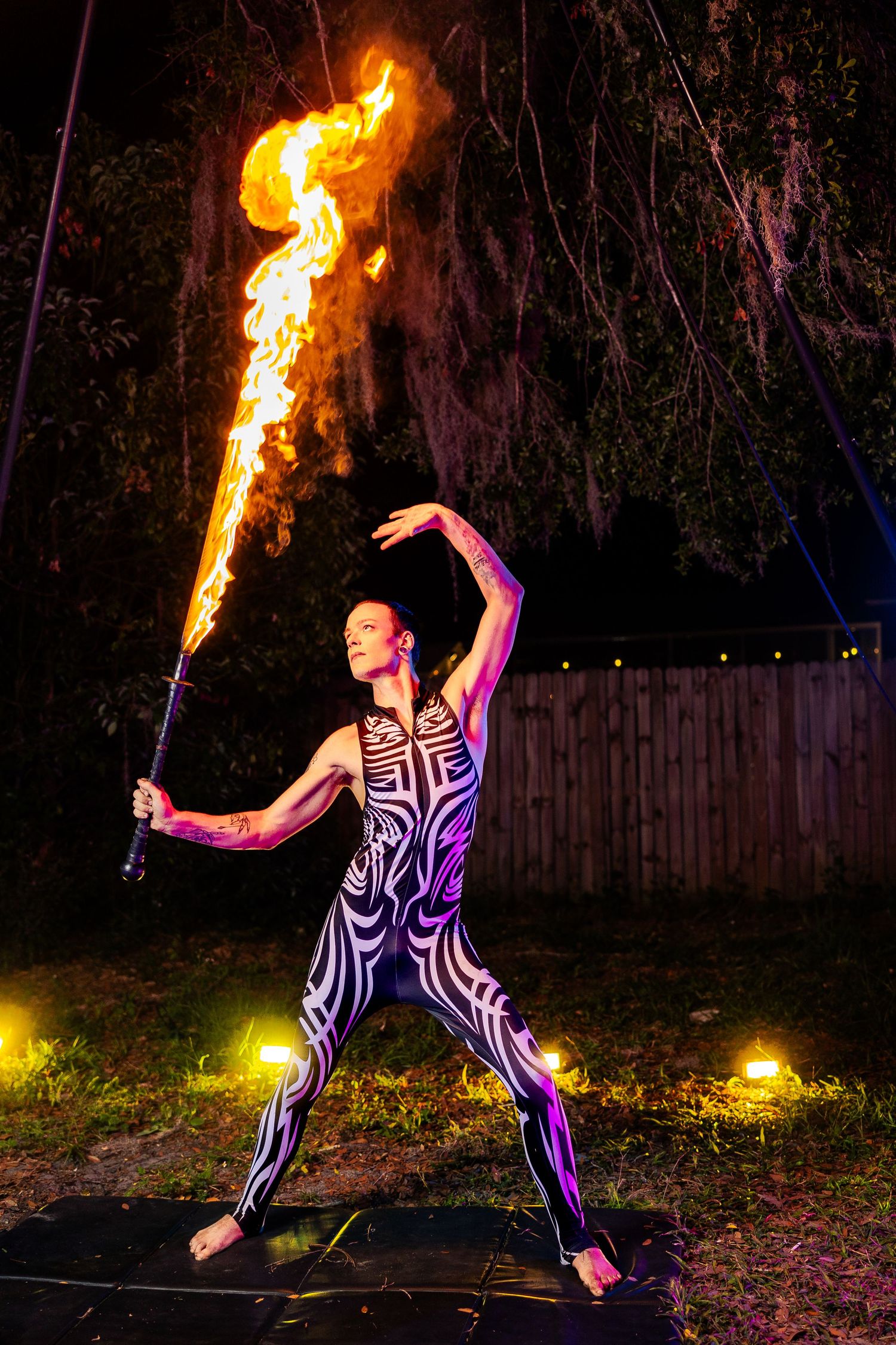 Fire dancer in black and white bodysuit performs with flaming staff outdoors at night.