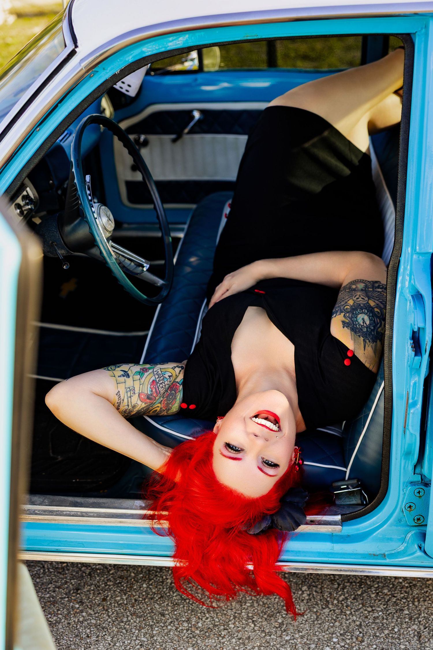 Tattooed woman with red hair lying back inside vintage blue classic car door.