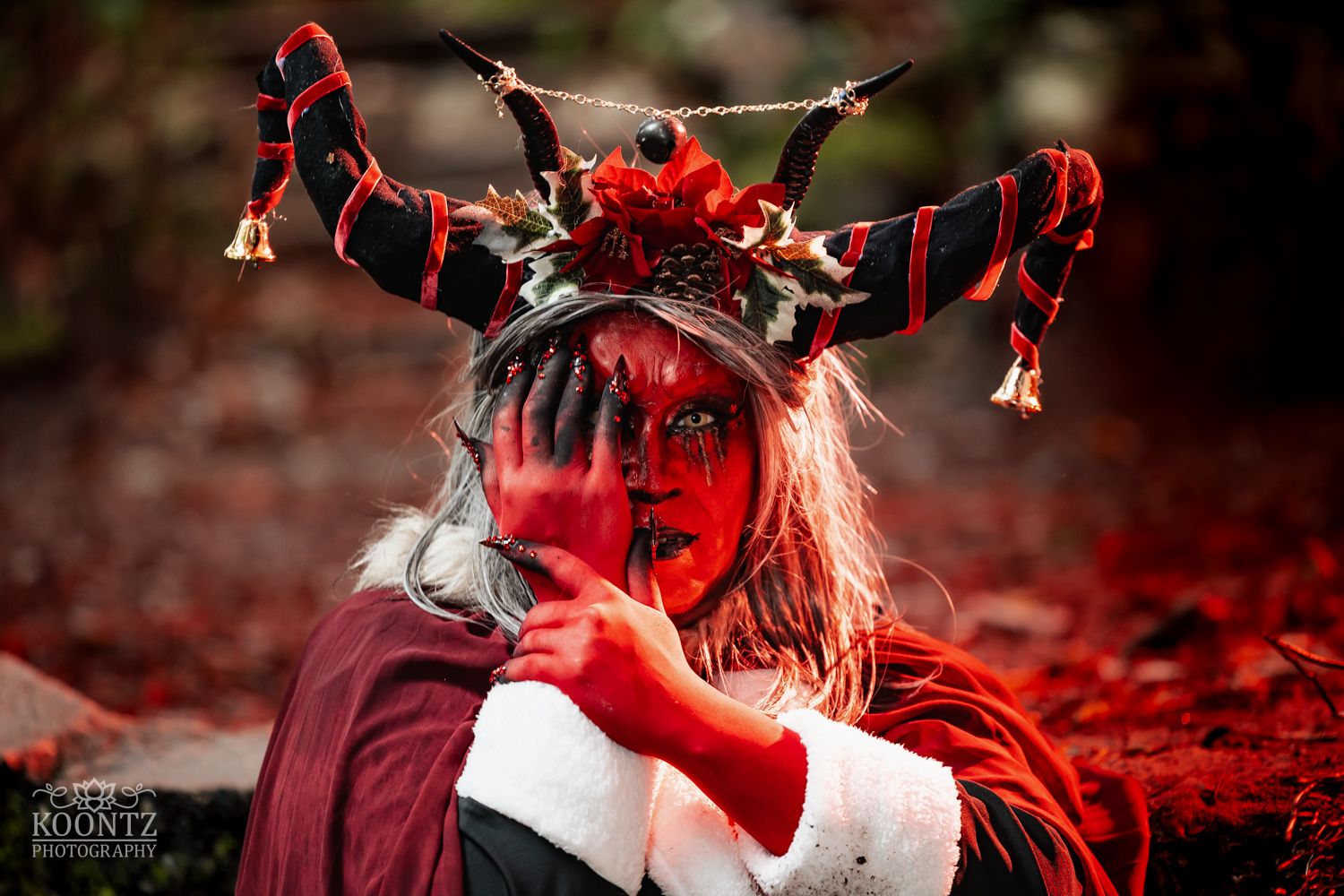 A cosplayer with red face paint and a floral antler headpiece peers over their arm in a red autumn forest.