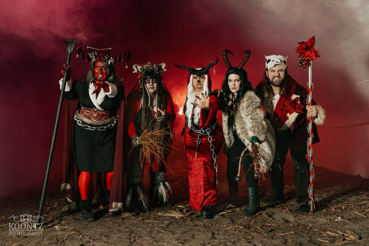 Five cosplayers in elaborate red and black devil and creature costumes pose in dramatic red smoke.