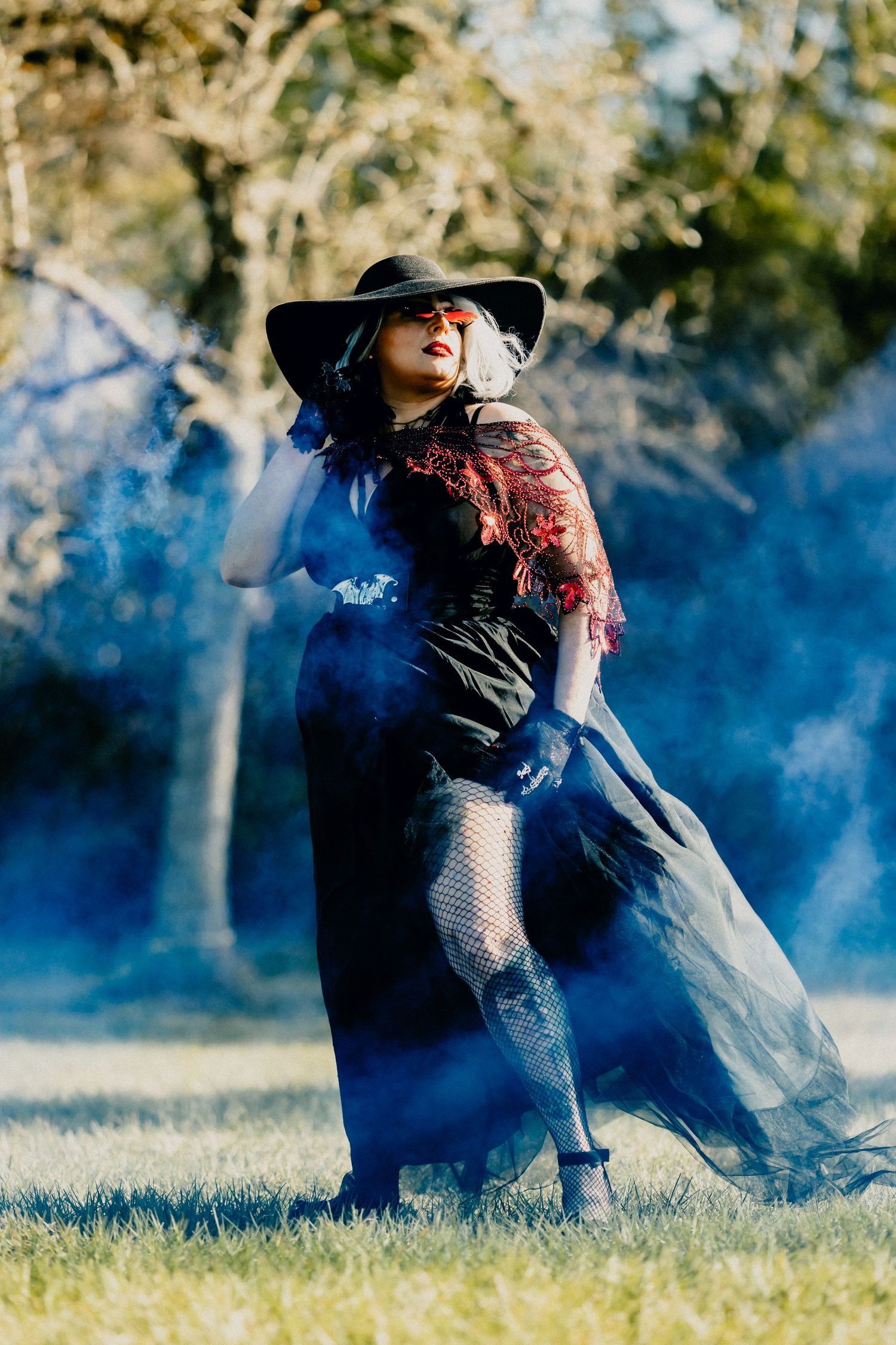 Woman in witch costume with black hat poses dramatically in blue smoke outdoors.