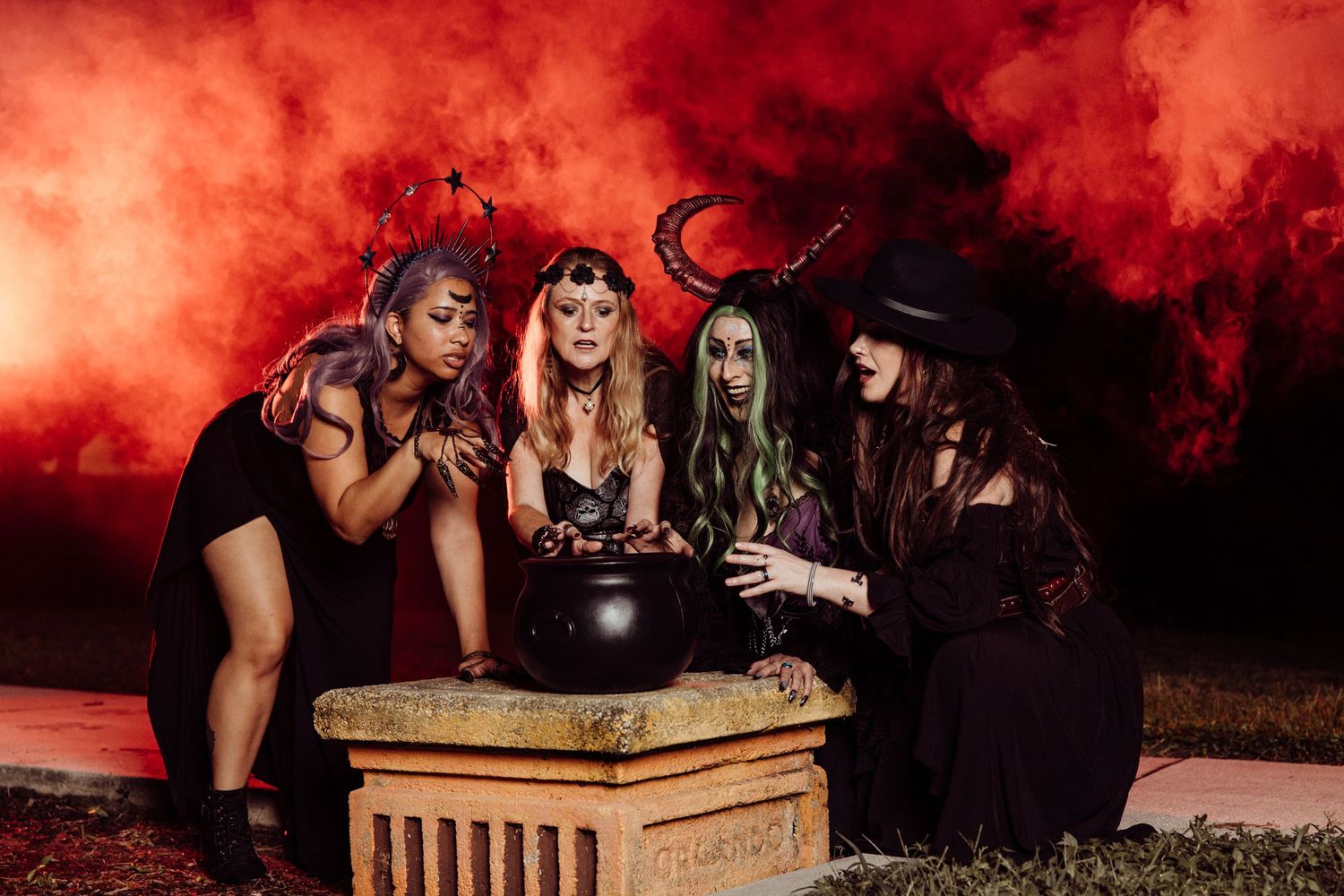 Four women in Halloween witch costumes gather around a cauldron with dramatic red smoke at night.