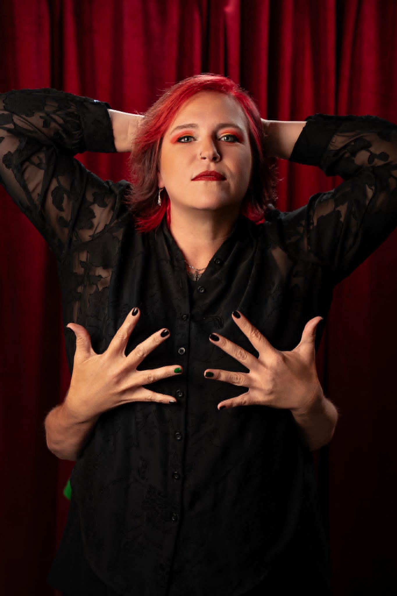 Woman with red hair in black lace shirt poses dramatically against red curtain backdrop with dark nail polish.