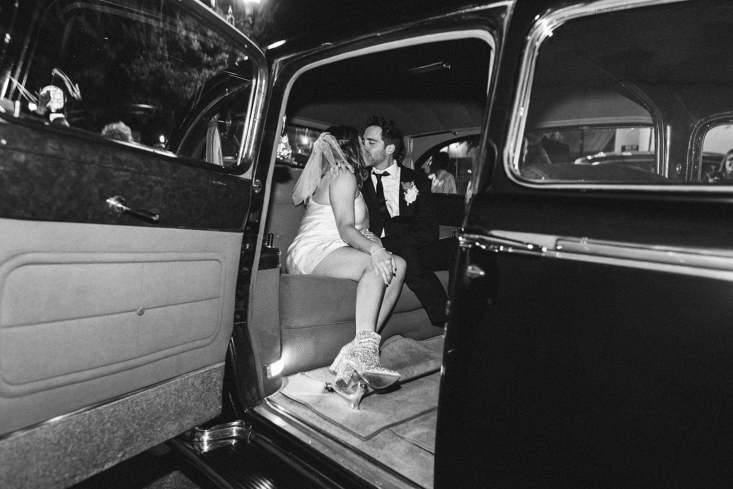 Black and white photo of bride and groom exiting a vintage classic car at night.