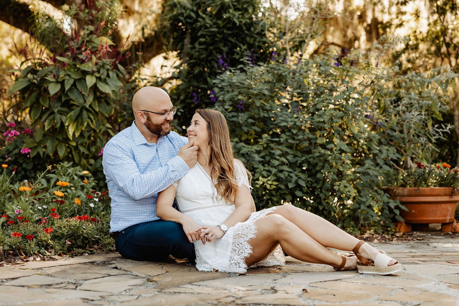 Couple sitting together in a lush garden, man in blue shirt touching woman's face in white lace dress.