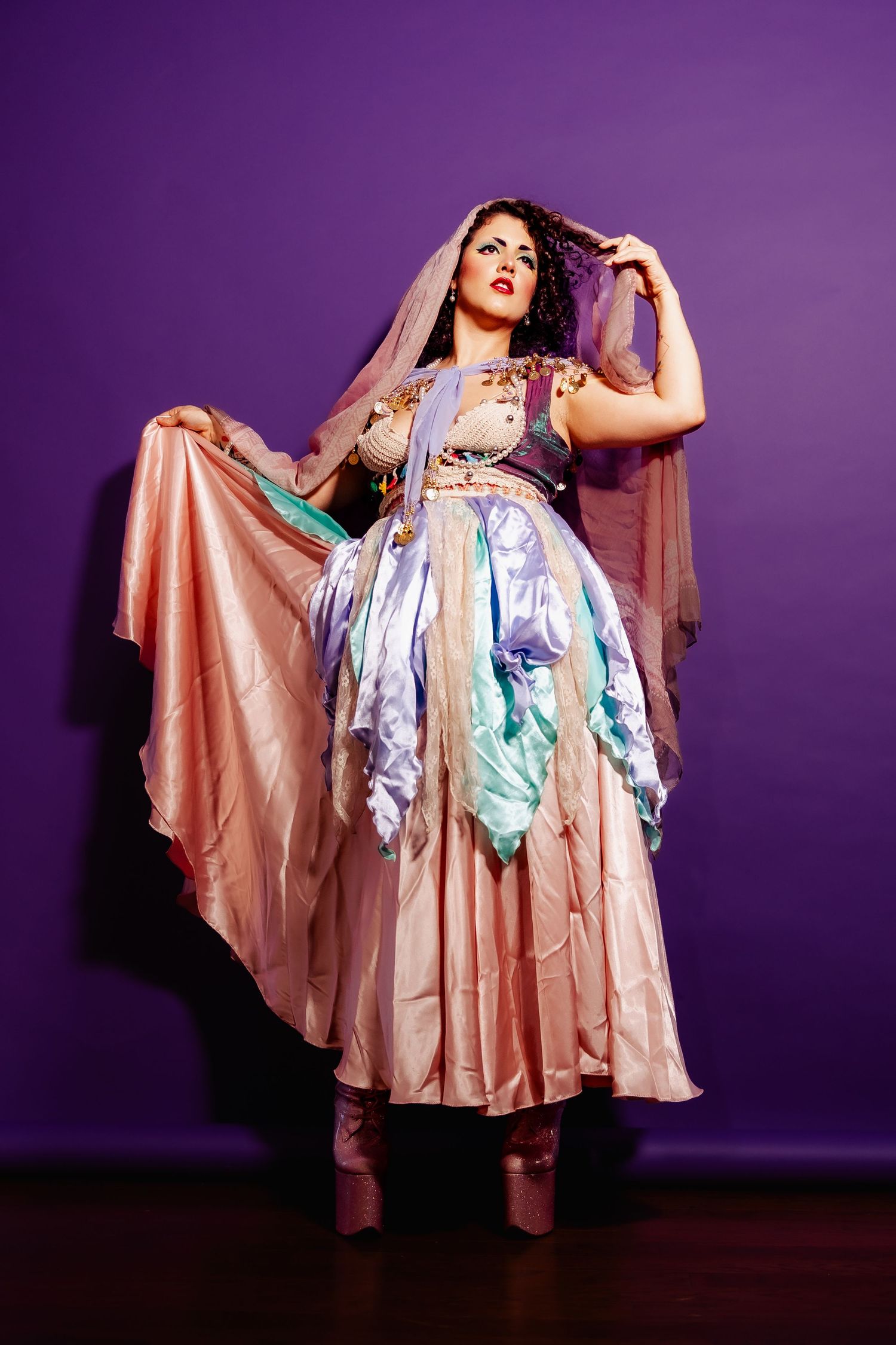 Woman in flowing pastel gypsy dress with lavender veil stands dramatically on purple background.