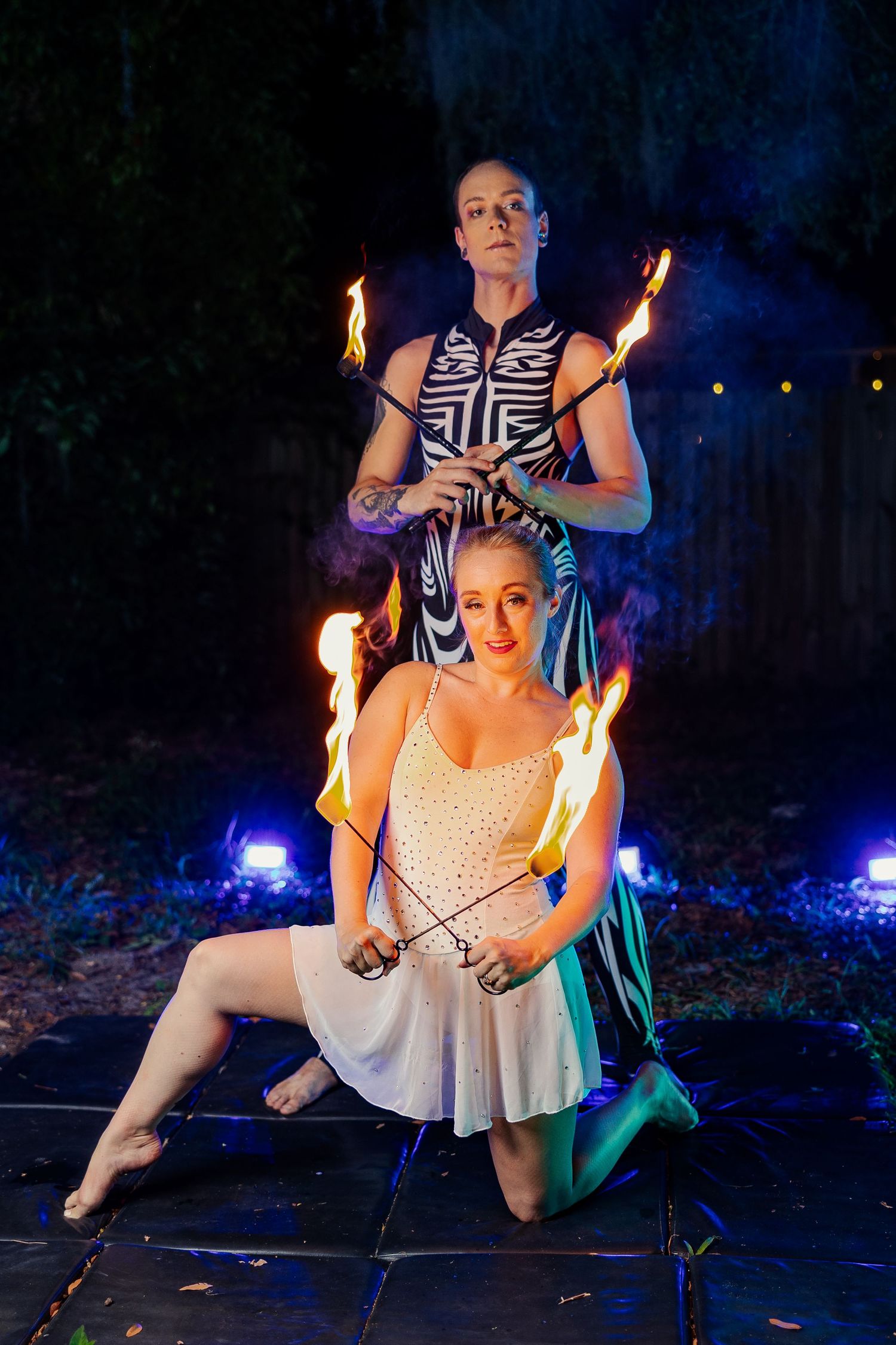 Fire performance duo poses with crossed flaming torches at night, woman in white dress kneeling before standing partner.
