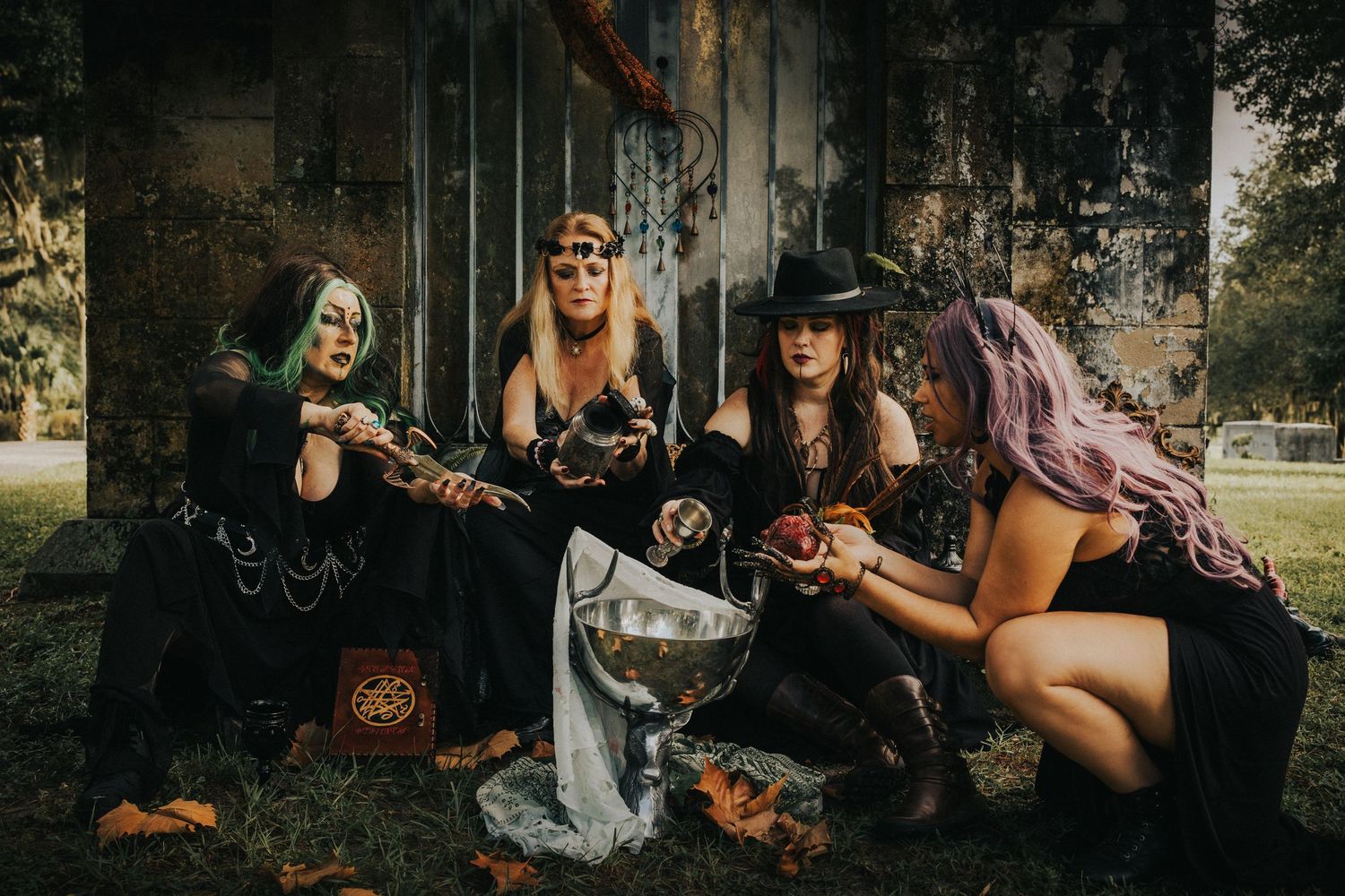 Four women dressed as witches sit together around a silver cauldron in an outdoor Halloween photoshoot.