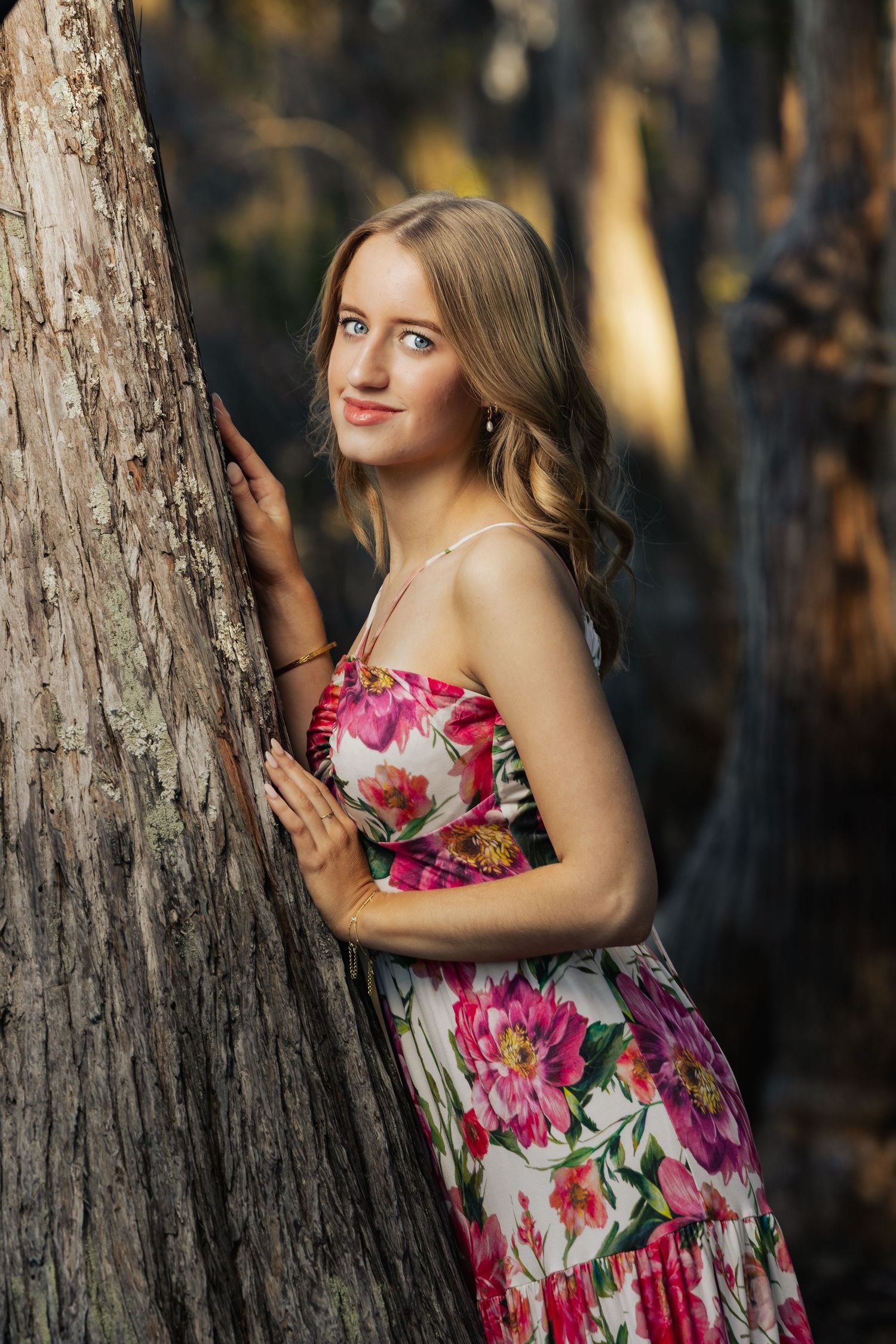 Young woman in floral dress leaning against a tree trunk in a sunlit forest setting.