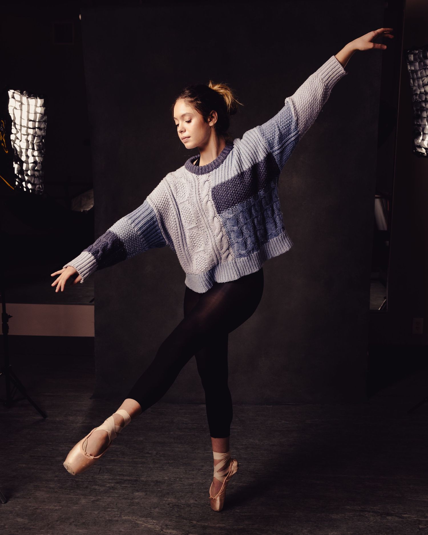 A ballet dancer in a blue sweater and black leggings poses gracefully on pointe shoes in a studio.